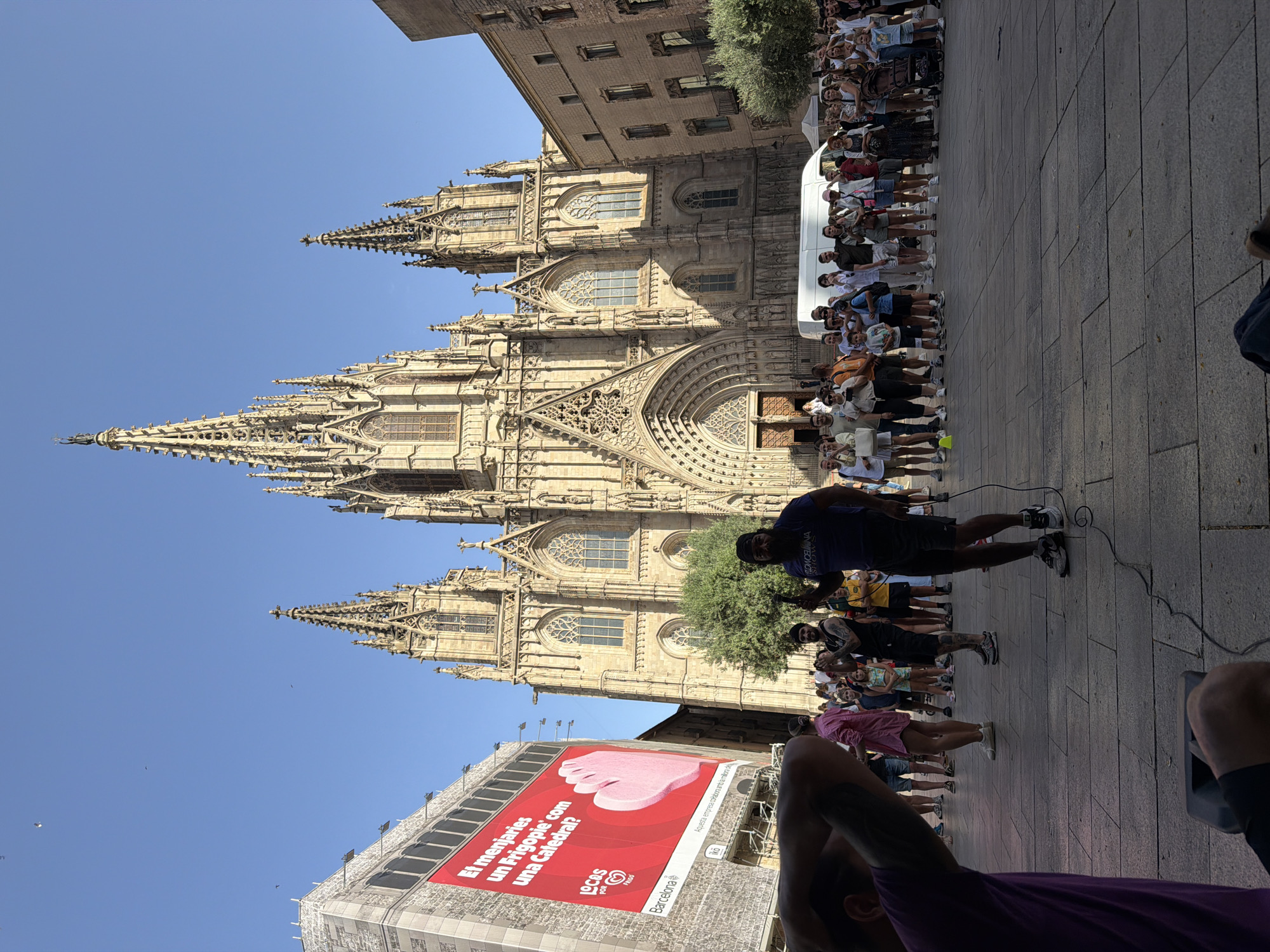 This is a photograph of Barcelona Cathedral in Barcelona, Spain. In the foreground, a man with a beard and a dark t-shirt is holding a microphone and speaking into it, with a cable running to a speaker on the ground. He is standing in a paved plaza, and a crowd of people are gathered behind him, watching. The cathedral, a large Gothic structure with spires and intricate stonework, dominates the background. To the left of the cathedral, a large building with a red billboard is visible. The sky is clear and blue.
