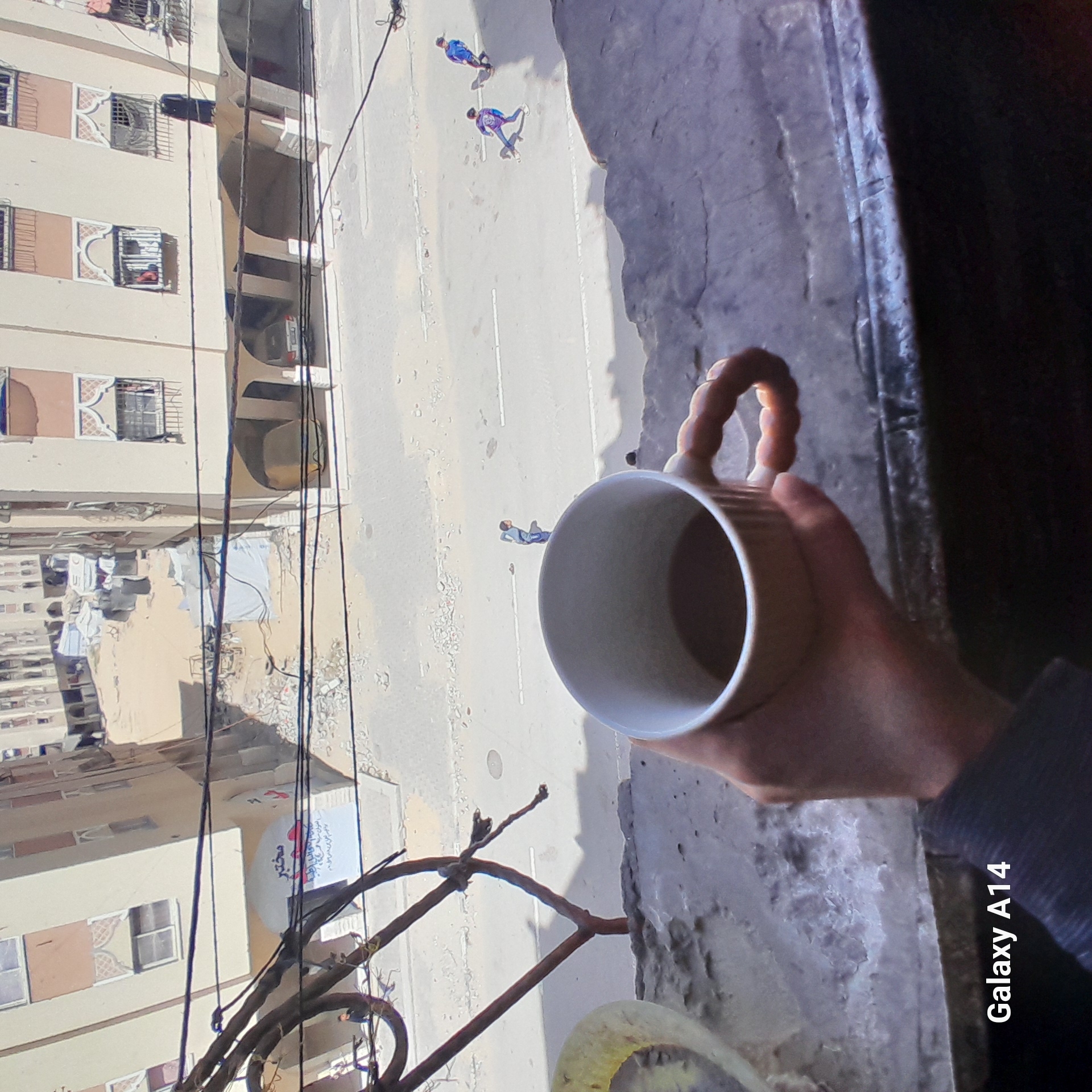 An overhead view captures a hand holding a ribbed white mug from a high vantage point, likely a balcony or window, looking down onto a street and adjacent buildings. The image is oriented sideways, with the street running vertically through the frame.
In the foreground, a person's right hand grasps a white ceramic mug with a distinctive ridged handle. The mug is empty and angled downwards. The hand and part of the arm are visible against a textured, dark grey surface, possibly a railing or ledge.
Below, a paved street stretches into the distance. Several people are visible on the street. Closer to the viewer, one person in light blue and white clothing stands. Further down the street, two figures in matching blue and purple outfits appear to be moving, possibly on skateboards or scooters, casting distinct shadows. White lines are painted on the asphalt road.
To the left of the street are several multi-story residential buildings, characterized by light-colored facades, numerous windows often fitted with metal grates, and balconies. One section shows clotheslines strung between buildings with laundry hanging to dry. An intricate pattern adorns the area around some windows. On one building, a circular sign is partially visible with Arabic text, including the word "مختبر" (Makhtabar, meaning "laboratory").
Numerous black electrical wires or cables crisscross the scene, connecting poles or buildings, suggesting an urban or semi-urban environment. The bright lighting indicates daytime, casting clear shadows. A "Galaxy A14" watermark is visible in the bottom right corner (when the image is rotated upright).