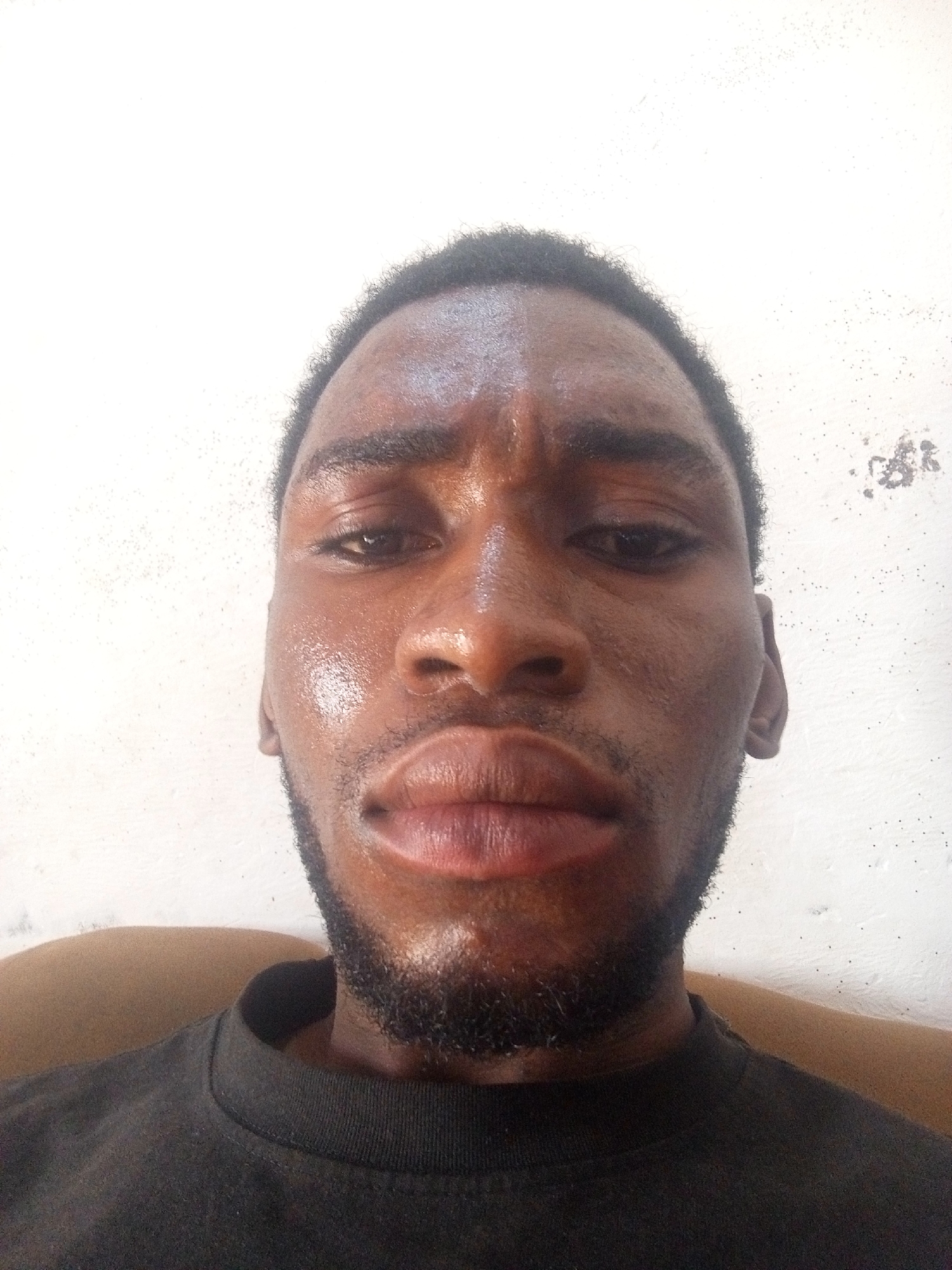 A close-up selfie of a dark-skinned man with a beard and short black hair. The man is sweating, particularly on his face. He is wearing a dark-colored shirt. The background is a plain white wall, suggesting an indoor location. It is impossible to confirm the location as Lokoja, Nigeria from the given image.