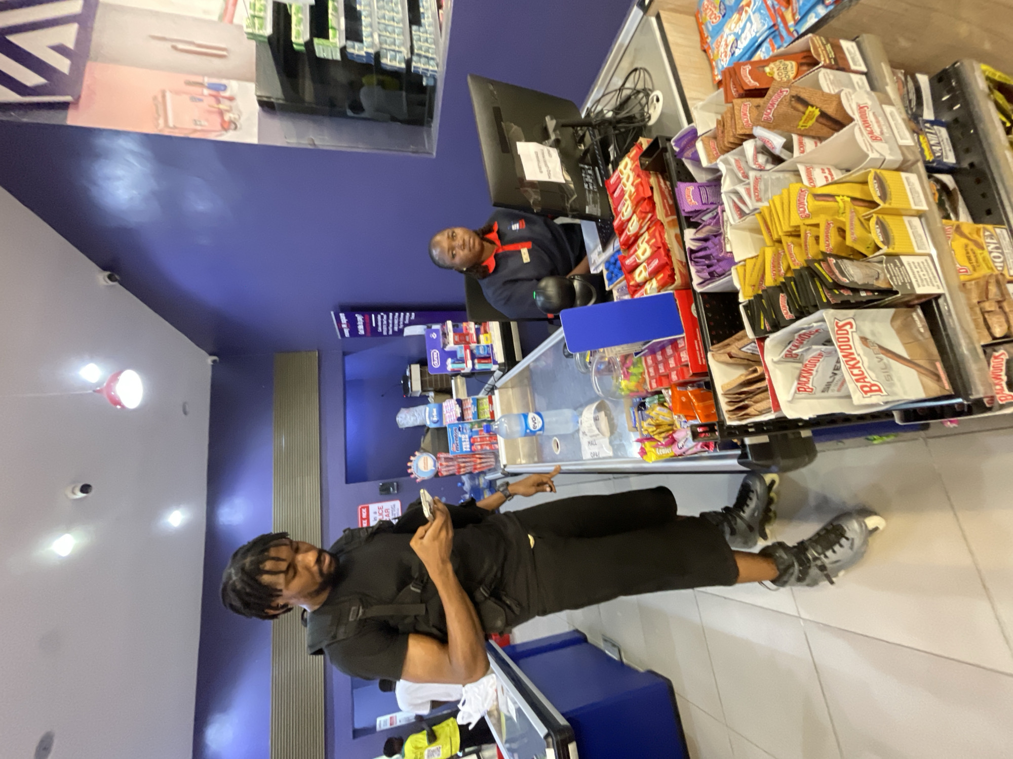 A Black male wearing a black t-shirt, black tactical vest, black shorts, and rollerblades is standing at a checkout counter in a brightly lit convenience store, looking at his phone. A Black female in a blue and red uniform with a name tag is seated behind the counter, operating a computer. Various products, including bottled water, candy, and cigarettes, are displayed on shelves and in bins around the counter. The store has purple walls and fluorescent lighting. The scene appears to be in Moba, Nigeria, given the context.