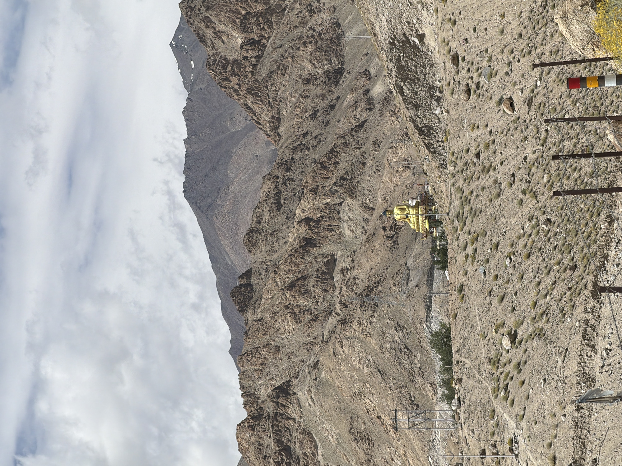 The image captures a wide shot of a mountainous landscape near Leh, India. In the mid-ground, a large golden Buddha statue is prominently displayed on a pedestal. The statue is seated and facing towards the viewer. The surrounding terrain is arid and rocky, with sparse vegetation. In the background, rugged mountains dominate the skyline, with some snow-capped peaks visible in the distance. The sky is overcast with scattered clouds. The foreground shows a barren, dusty ground with a fence constructed of barbed wire and posts. Electrical poles are also visible in the mid-ground. The overall scene conveys a sense of remoteness and spiritual significance.