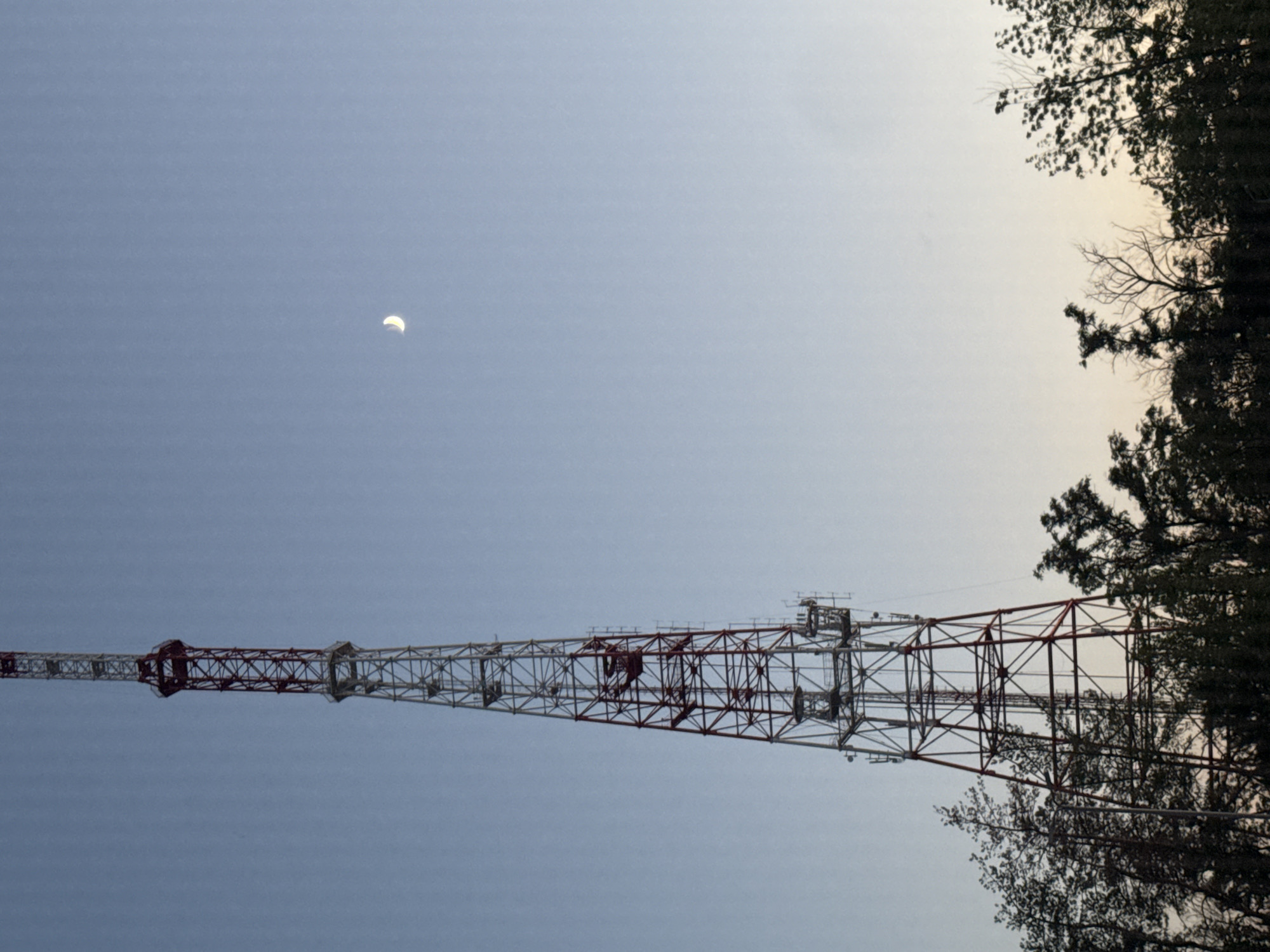 A tall, red and white lattice tower stands prominently in the center of the frame, reaching towards a pale blue sky. The tower has several platforms and antennae. In the upper right quadrant of the sky, a slender crescent moon is visible. The lower portion of the image is filled with the dark silhouettes of trees and foliage. The scene appears to be set in the city of Mariupol, Ukraine, likely during the evening or early morning given the sky conditions. There are no people or animals present in the image.