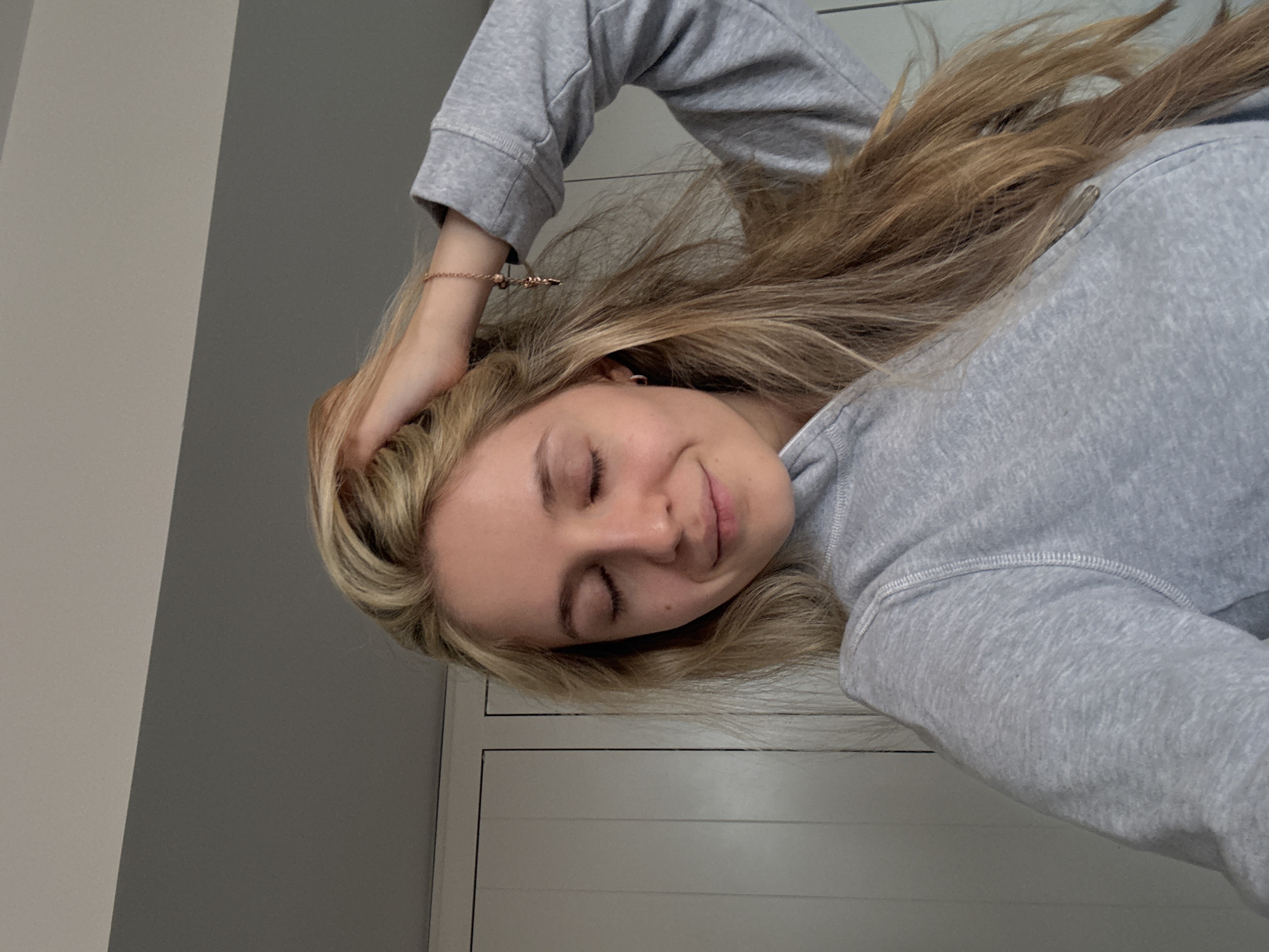 A young woman with blonde hair is wearing a gray sweatshirt and is lying down, with her eyes closed. Her right hand rests on her head, and a bracelet is visible on her wrist. The background is a combination of light gray wall and white paneling. The location is not discernible from the image provided.