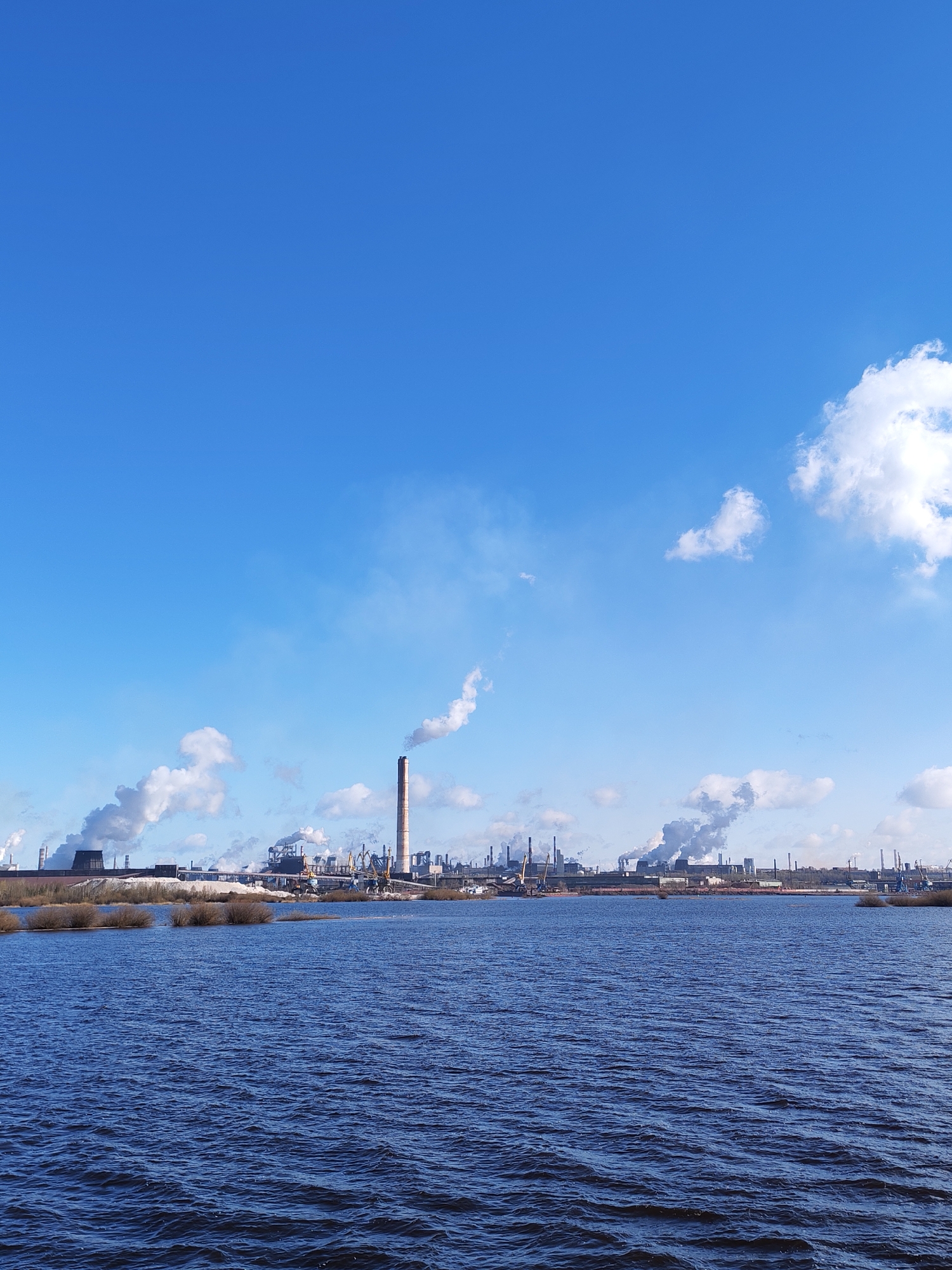 The image shows a body of water in the foreground with a large industrial complex in the background. The sky is blue with scattered white clouds. Several smokestacks are visible, emitting plumes of white smoke. The water is dark blue with ripples. There are some patches of vegetation near the water's edge. The location is in the city of Unknown, Unknown.