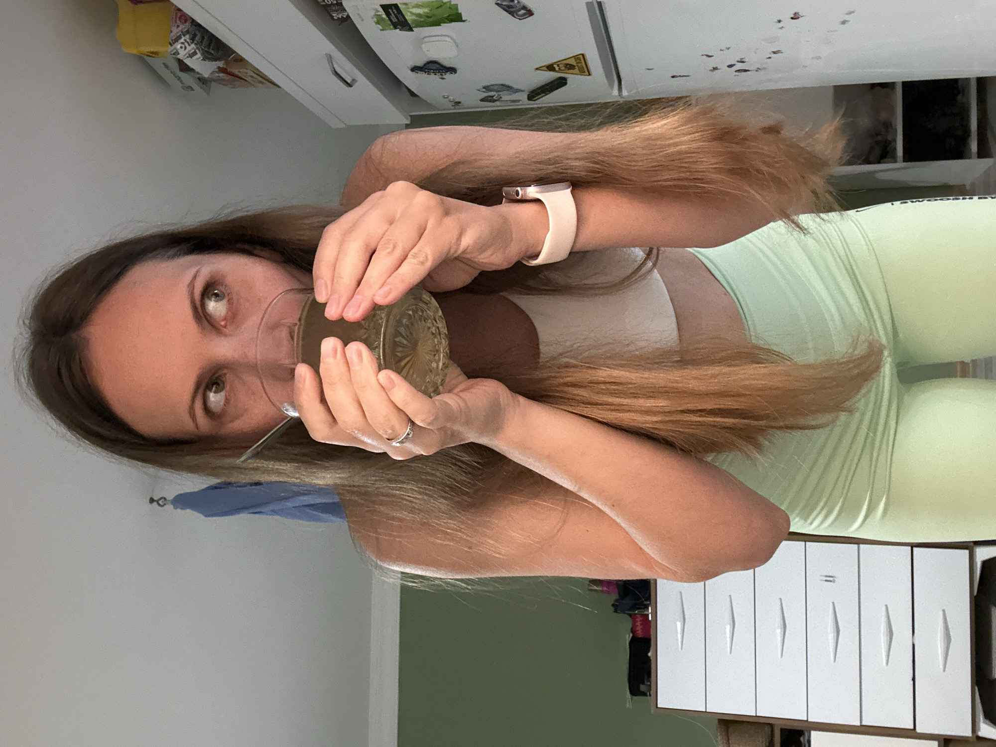 A person with long, brown hair is drinking from a glass with a spoon in it. They are wearing a white top and light green leggings. They are wearing a pink wristwatch. They are standing in a room with white walls, a white cabinet, and a green wall. The location is unknown, but based on the prompt, it is presumed to be Fethiye, Turkey.