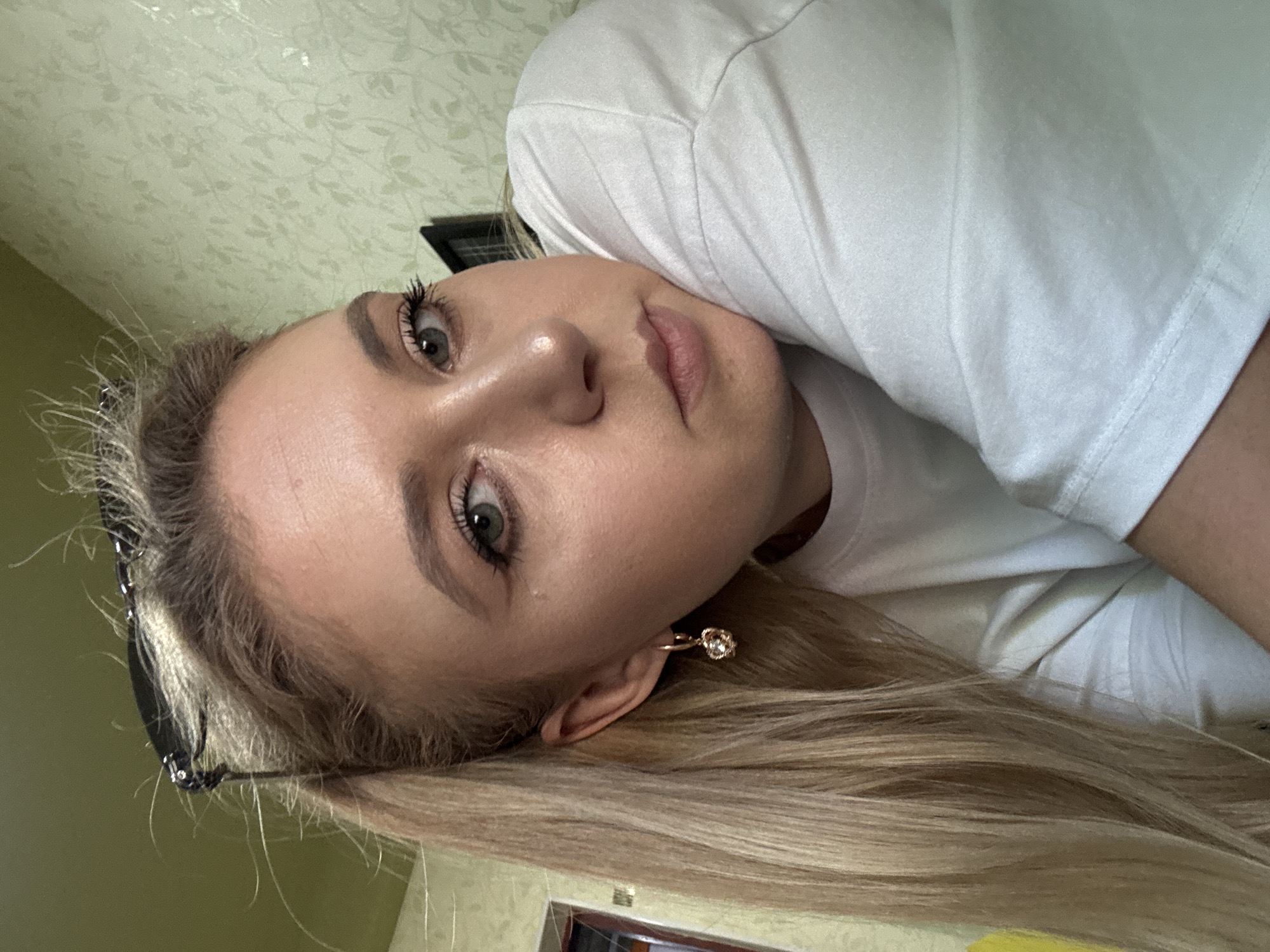 A blonde woman wearing a white t-shirt and sunglasses on her head is pictured in a close-up shot. She has green eyes and is wearing a heart-shaped earring. The background appears to be a room with light green floral wallpaper. The lighting suggests it is daytime.