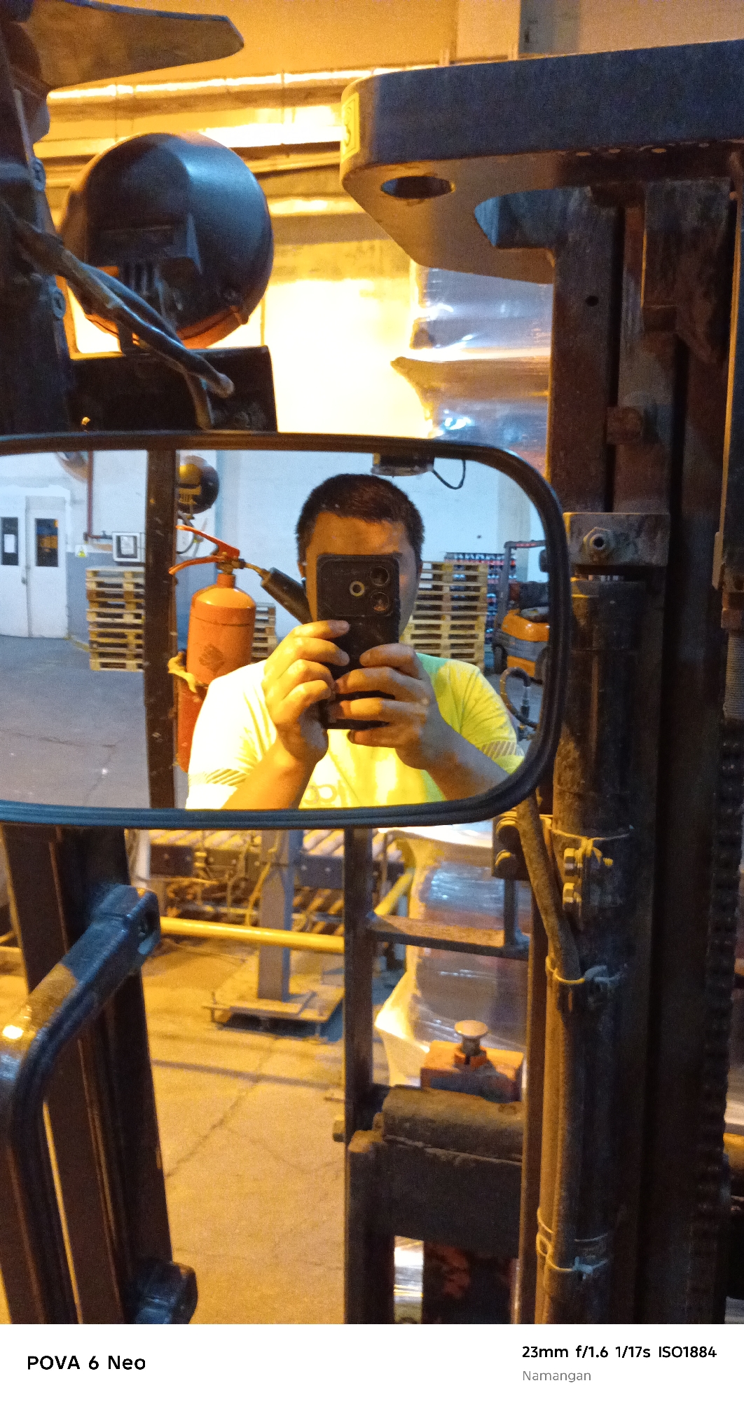The image shows a reflection in a side-view mirror of a forklift. The reflection captures a person taking a selfie with a smartphone. The person is wearing a bright yellow t-shirt and is holding a black smartphone in front of their face, with the camera facing the mirror. In the background of the reflection, a red fire extinguisher is visible mounted on the forklift. The environment appears to be a warehouse or industrial setting, with stacks of pallets and other equipment. The bottom of the image contains text indicating camera settings and the location "Namangan".