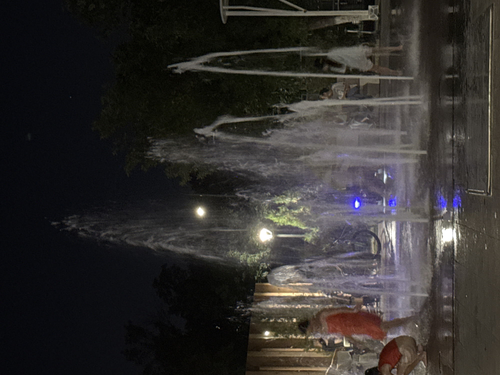 The image depicts an outdoor fountain area at night, presumably in Shymkent, Kazakhstan, as indicated by the prompt. Several individuals are present. In the foreground, on the left, a person wearing a red top and light-colored shorts is crouching, with their feet in the shallow water. To their right and slightly further back, a person in a red dress stands. In the background, several other individuals are visible, some sitting and some standing near the fountain. The fountain itself consists of multiple vertical jets of water spraying upwards and outwards, creating a misty atmosphere. The ground around the fountain is wet and reflective due to the water. Trees and some architectural elements, including lighting fixtures and a wooden fence, are visible in the background. The overall scene suggests a public recreational space at night.
