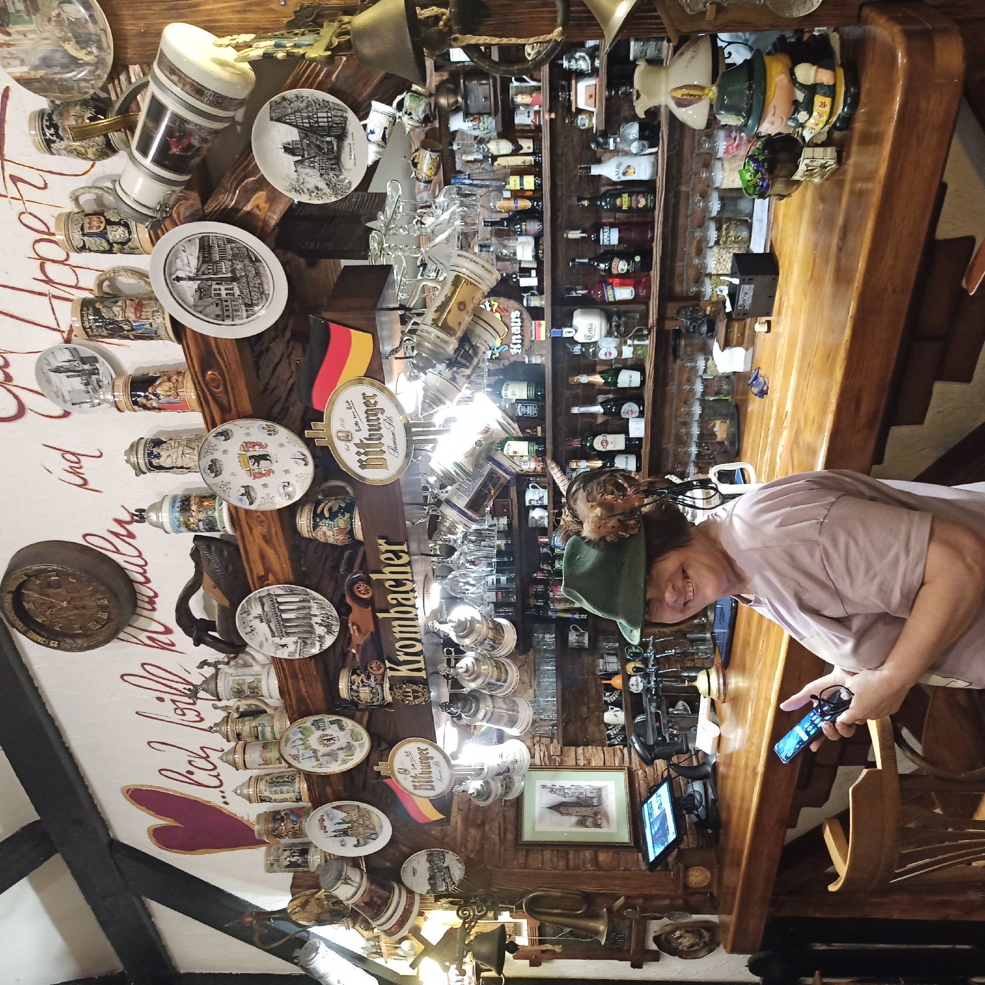 The image depicts the interior of a Bavarian-themed bar, likely located in Chernivtsi, Ukraine. The walls are decorated with German plates, steins, and other memorabilia, including a large wooden clock and a painted heart. A woman wearing a green hat with feathers is standing behind the wooden bar, holding a mobile phone and a pair of glasses. She is smiling at the camera. The bar itself is stocked with various bottles of alcohol, glassware, and beer taps. A digital tablet is visible on the bar counter. The overall atmosphere suggests a cozy and traditional establishment.