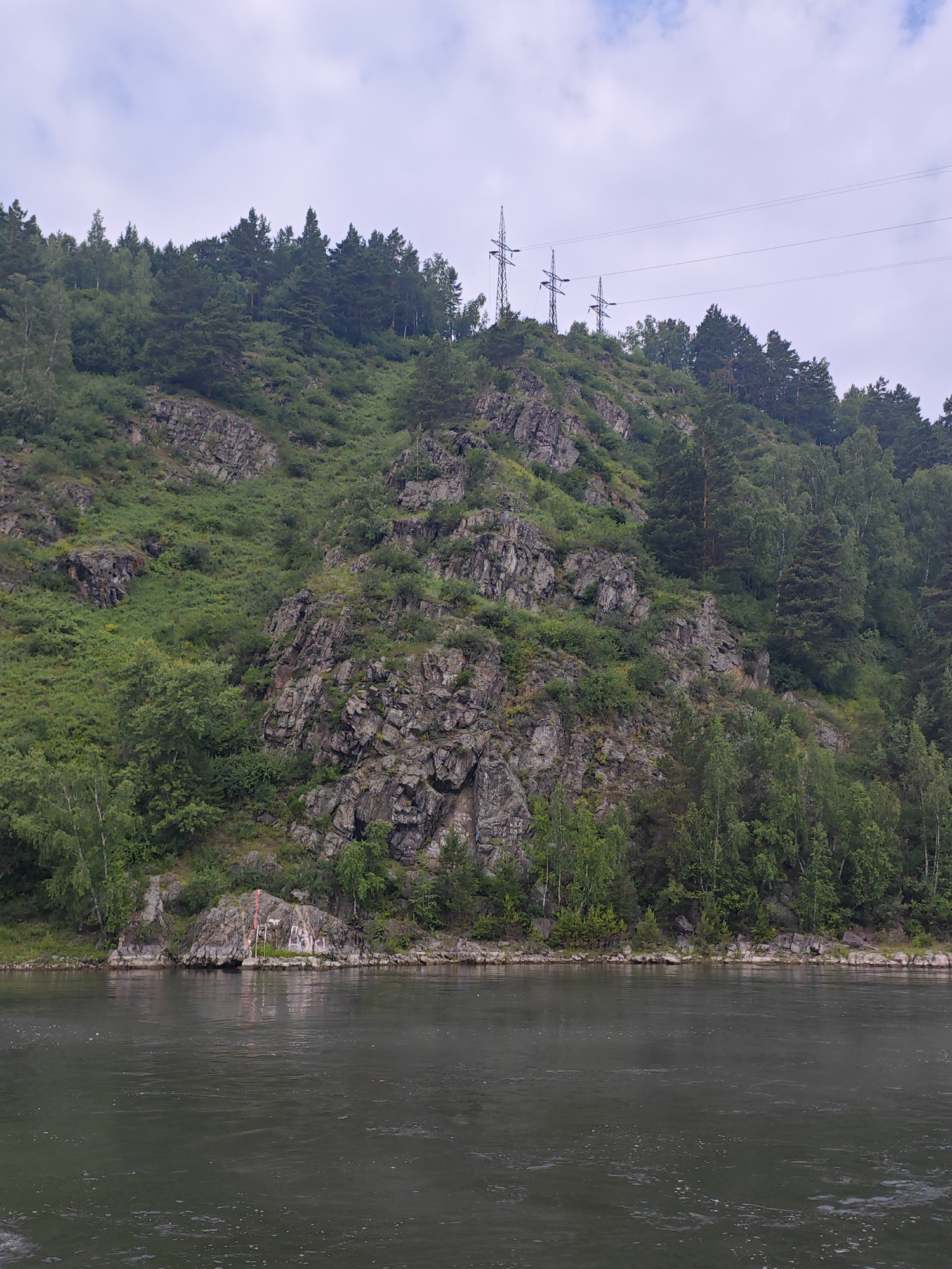 A wide, dark gray river flows in the foreground, with ripples and some white foam visible on its surface. On the right bank of the river is a steep, rocky hill covered in dense green vegetation and trees. The rocks are gray and jagged. At the top of the hill, several tall, thin power line towers with wires stretching between them are visible against a cloudy, pale sky. Some lighter green trees are scattered among the darker green foliage on the hillside. In the lower left corner of the image, on the bank of the river, there is a rocky outcrop with some graffiti or markings on it and a red and white striped marker pole.