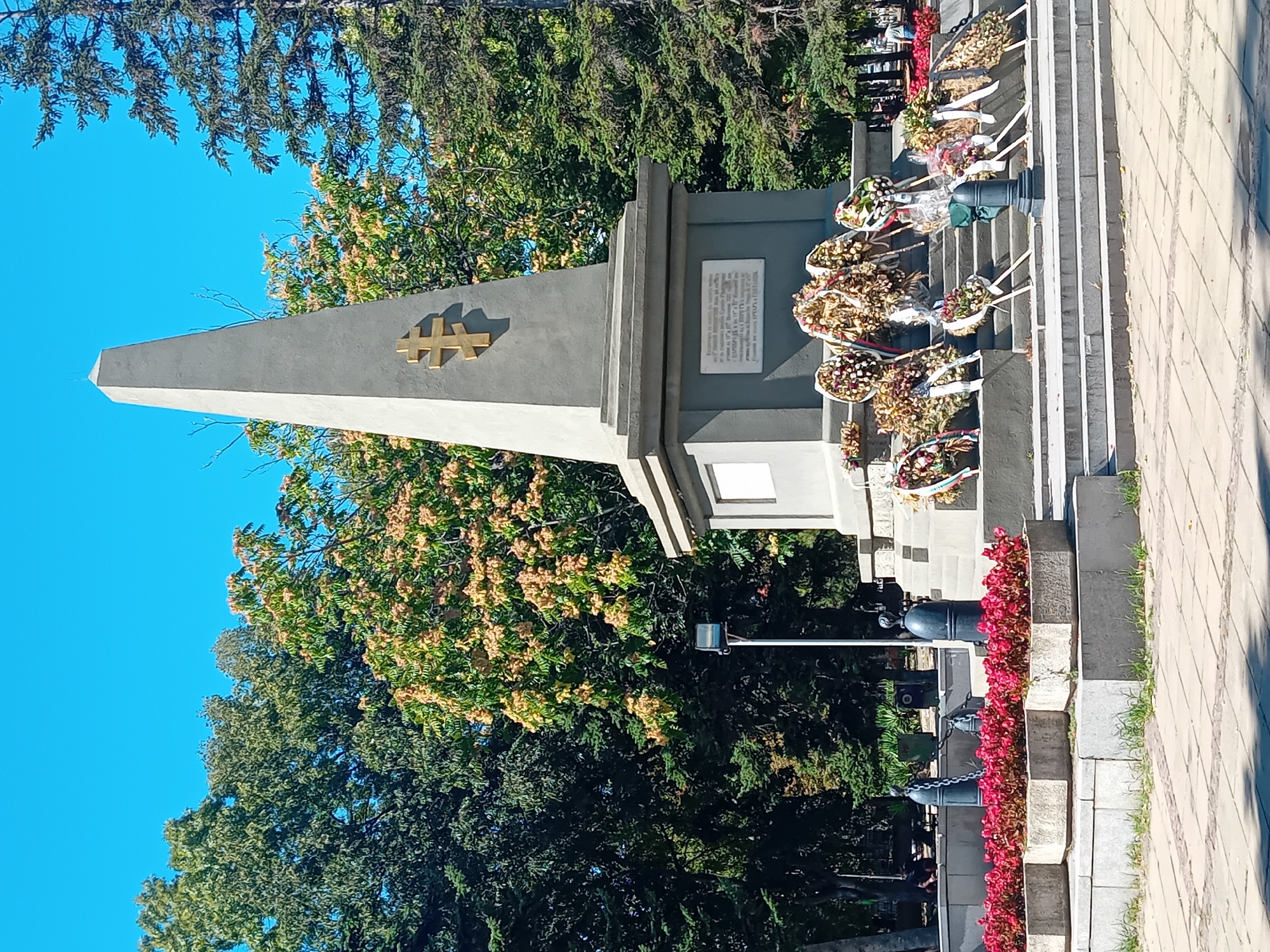 The image depicts the Monument to the Bulgarian Aviation, located in Varna, Bulgaria. The monument is an obelisk made of concrete with a golden double-barred cross on its facade. Surrounding the monument are several wreaths of flowers, suggesting a recent commemorative event. In the foreground, there are planter boxes filled with red flowers. The monument is situated in an outdoor area with trees and a clear blue sky. Some people can be seen in the background, near the monument and in the surrounding park.