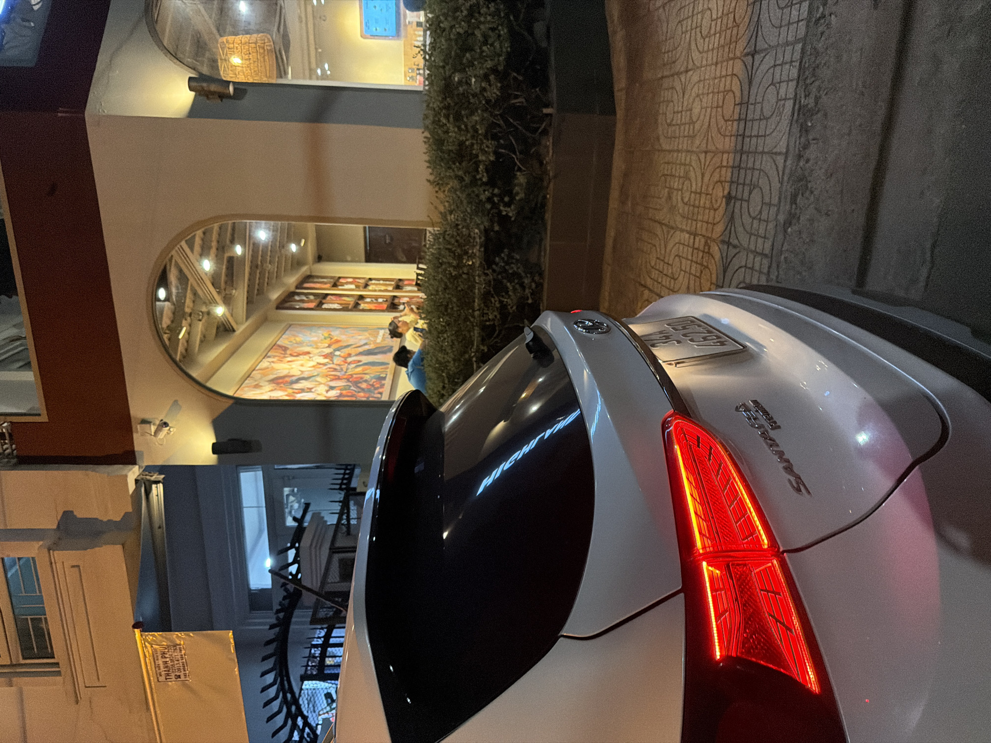 The image shows the rear of a white Hyundai Santa Fe parked on a sidewalk in Nha Trang Ward, Vietnam. The car is positioned on a patterned concrete sidewalk. In the background, there's a building with an arched entrance to what appears to be a cafe or restaurant. Inside, several people are visible sitting at tables. The building has decorative lighting and artwork on the walls. A street lamp is visible on the side of the building. A yellow banner with Vietnamese text is hanging on the building to the left. The scene appears to be at dusk or night, with artificial lighting illuminating the area.