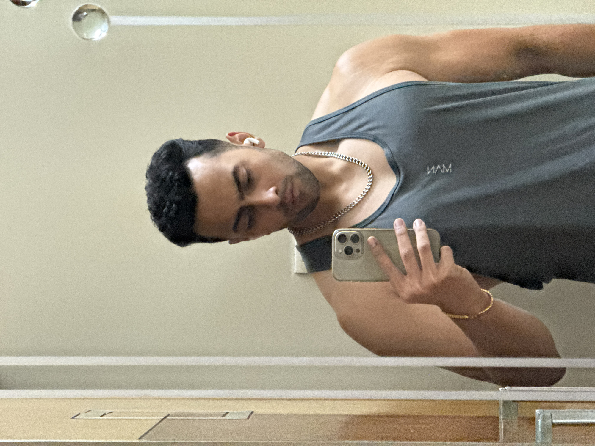 A man wearing a grey tank top and gold jewelry, including a necklace and bracelet, is taking a selfie in a mirror. He is holding a phone with a gold-colored case. He has an earbud in his left ear. The background appears to be a bathroom or dressing area. The location is undetermined.
