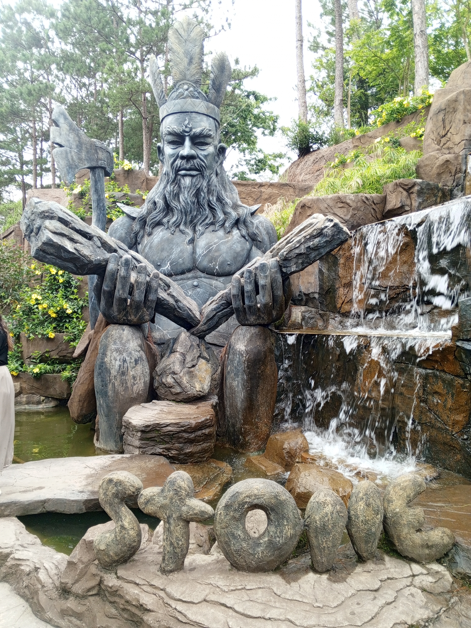 The image depicts a large, imposing statue of a warrior with a long beard and elaborate headdress, holding two large logs. The statue is seated in front of a rock formation with a waterfall cascading down. In the foreground, stone letters spell out "STONE". The setting appears to be a park or garden, with lush greenery and trees in the background. This scene is located at the Datanla Waterfall amusement park in Da Lat, Vietnam, specifically near the roller coaster attraction.