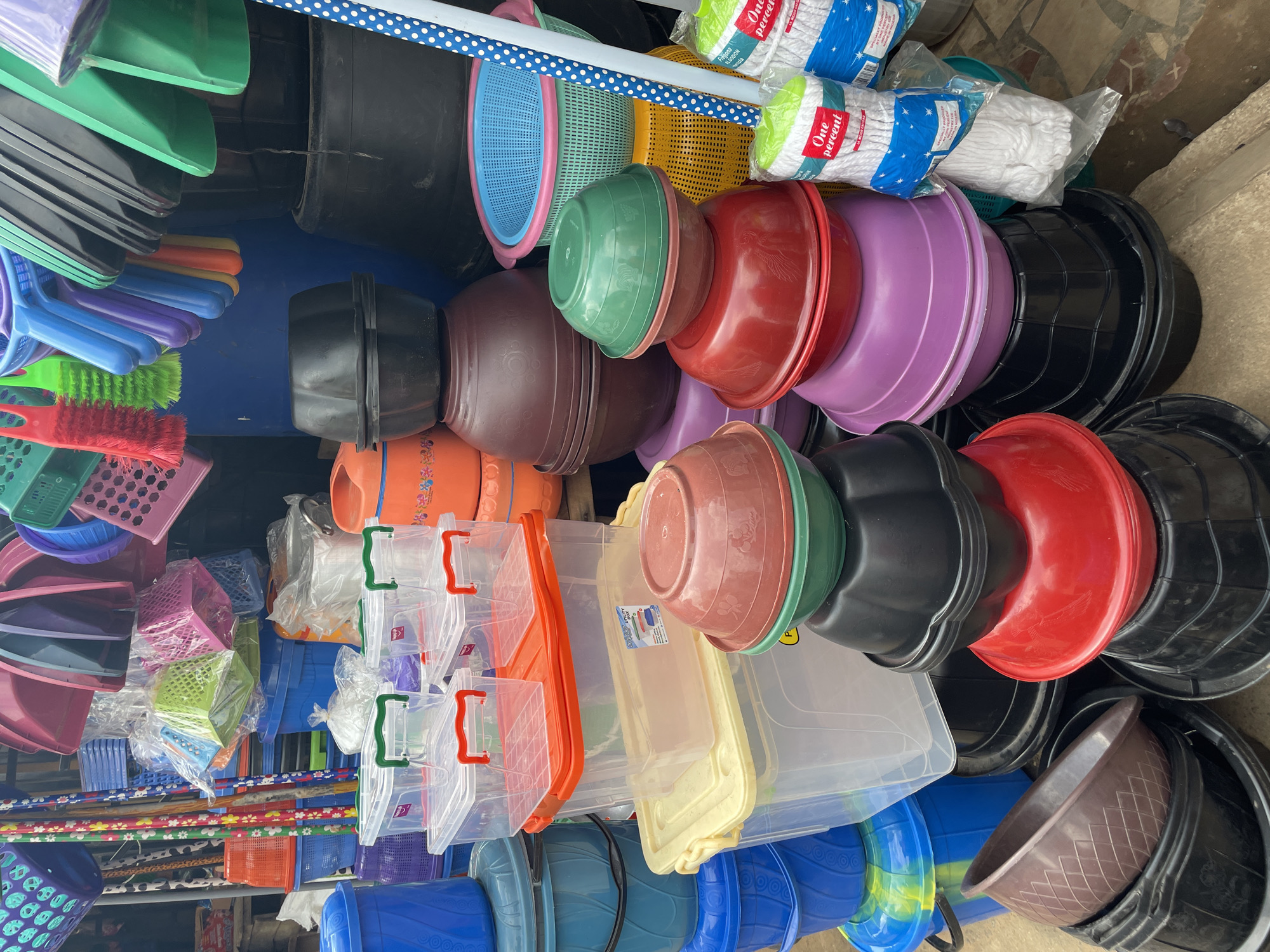 This image depicts an outdoor market stall in Akwanga, Nigeria, displaying a variety of plastic household goods. The stall is filled with stacks of colorful plastic basins, bowls, buckets, and storage containers in various sizes and shades of blue, red, green, purple, black, and brown. Among the larger items, there are several clear plastic storage boxes with red and green lids. Also visible are brushes with red and green bristles, and a mop with a white head and a blue and white patterned handle. In the background, more plastic items and what appear to be brooms or long-handled tools are stacked. The ground appears to be concrete. There are no people or animals visible in the frame, and no overt actions or interactions are taking place. The overall scene is one of organized retail display within a market setting.