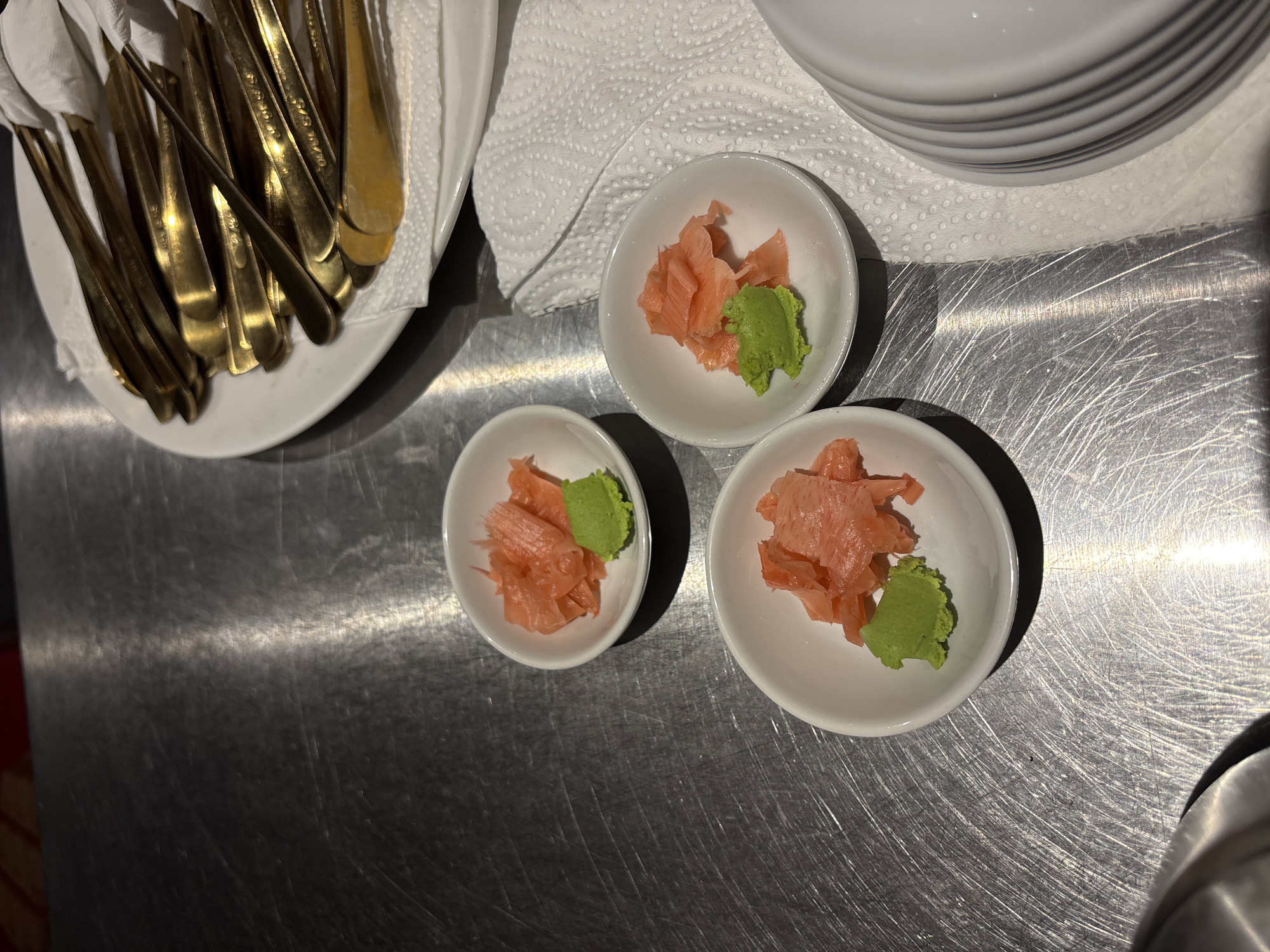 The image shows three small white bowls containing pickled ginger and wasabi on a brushed metal surface. In the background, a white plate is filled with gold-colored cutlery wrapped in white napkins. The scene appears to be in Kuta Selatan, Indonesia, likely in a restaurant setting.