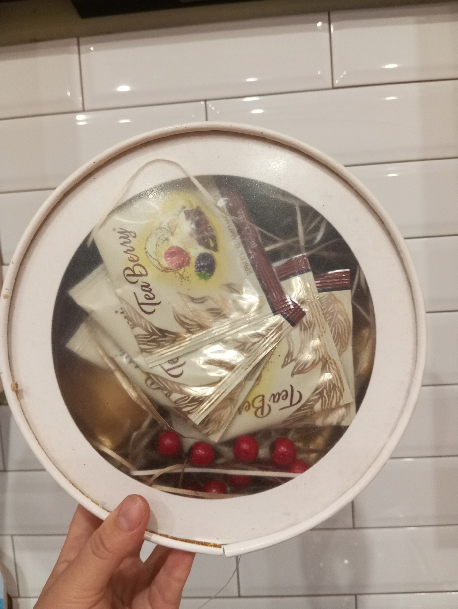 A person's hand is holding a round, white tin containing several individually wrapped tea bags. The tea bags are golden in color and have "Tea Berry" printed on them in an elegant font, along with an illustration of berries. There are also some small, red, round objects at the bottom of the tin, possibly candies or decorative elements. The tin is placed on a surface with a white tiled background. The lighting suggests an indoor setting. The image does not provide any information about the city of Verkhnyaya Pyshma, Russia.
