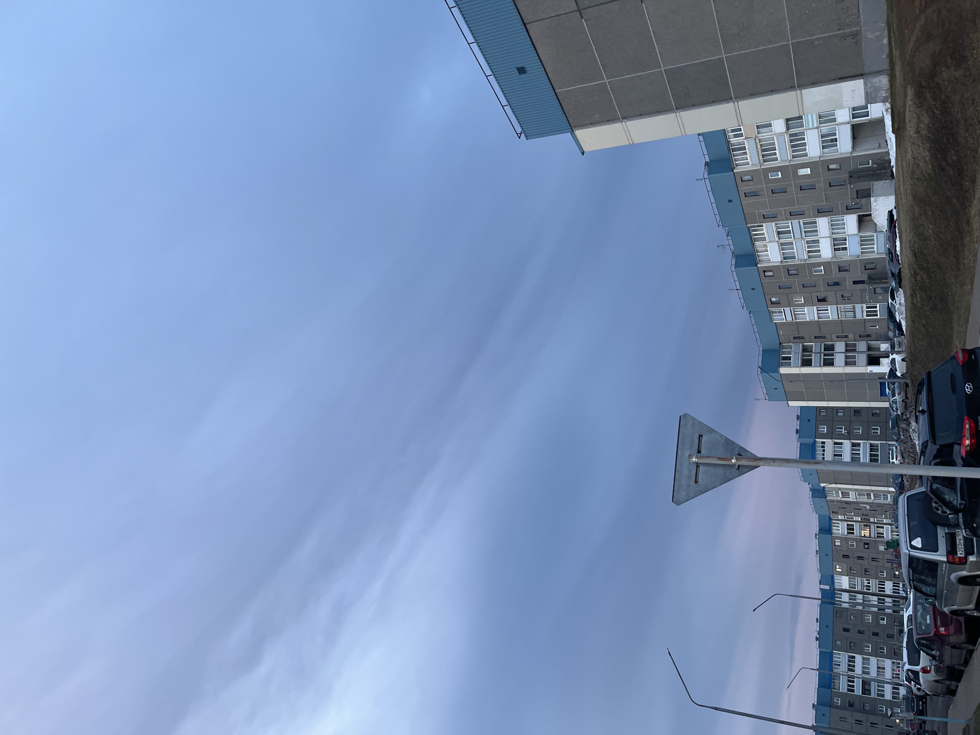 The image shows a view looking up at a sky that is mostly blue with light streaks of clouds. The tops of buildings are visible on the right side and top of the frame. A signpost is visible near the middle of the frame, oriented vertically. Cars are parked along the edge of the building to the right. Based on the location identified in the prompt, this is likely a residential area in the city of Марковский, Russia.