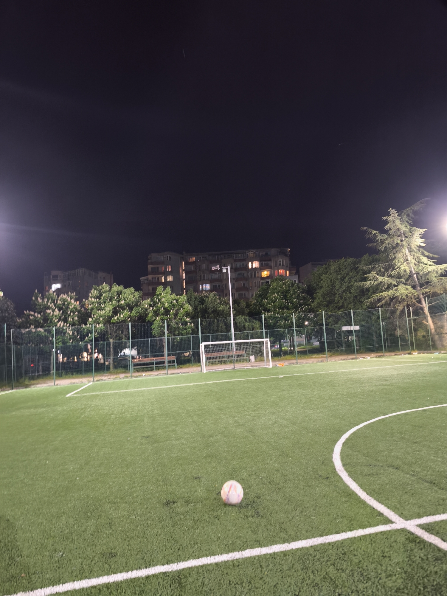 A soccer ball rests on the edge of a soccer field's center circle on a dark night. The field is surrounded by a fence and trees. Beyond the fence are buildings with lit windows. The location is in Burgas, Bulgaria.