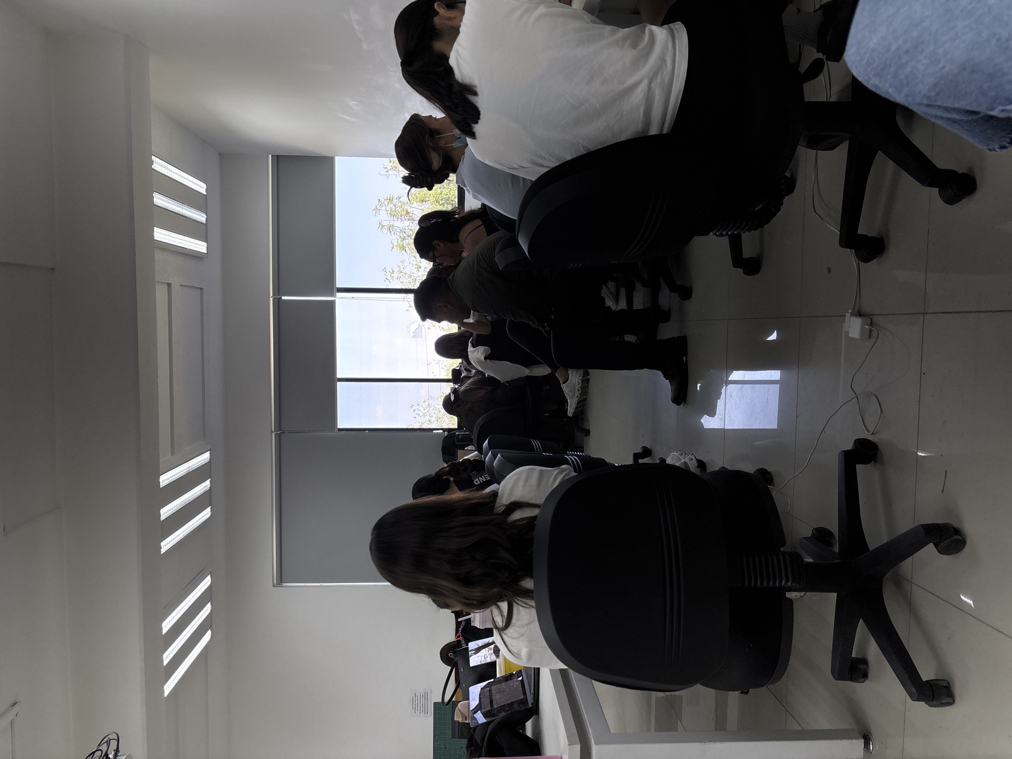 The image shows a classroom setting at a university in San Andrés Cholula, Mexico. There are several students sitting at desks, appearing to be engaged in individual work on laptops or tablets. The room is well-lit with natural light coming through the windows and supplemented by fluorescent lights on the ceiling. The students are seated in black office chairs. Some are wearing masks, while others are not. The classroom environment suggests a focused and academic setting.