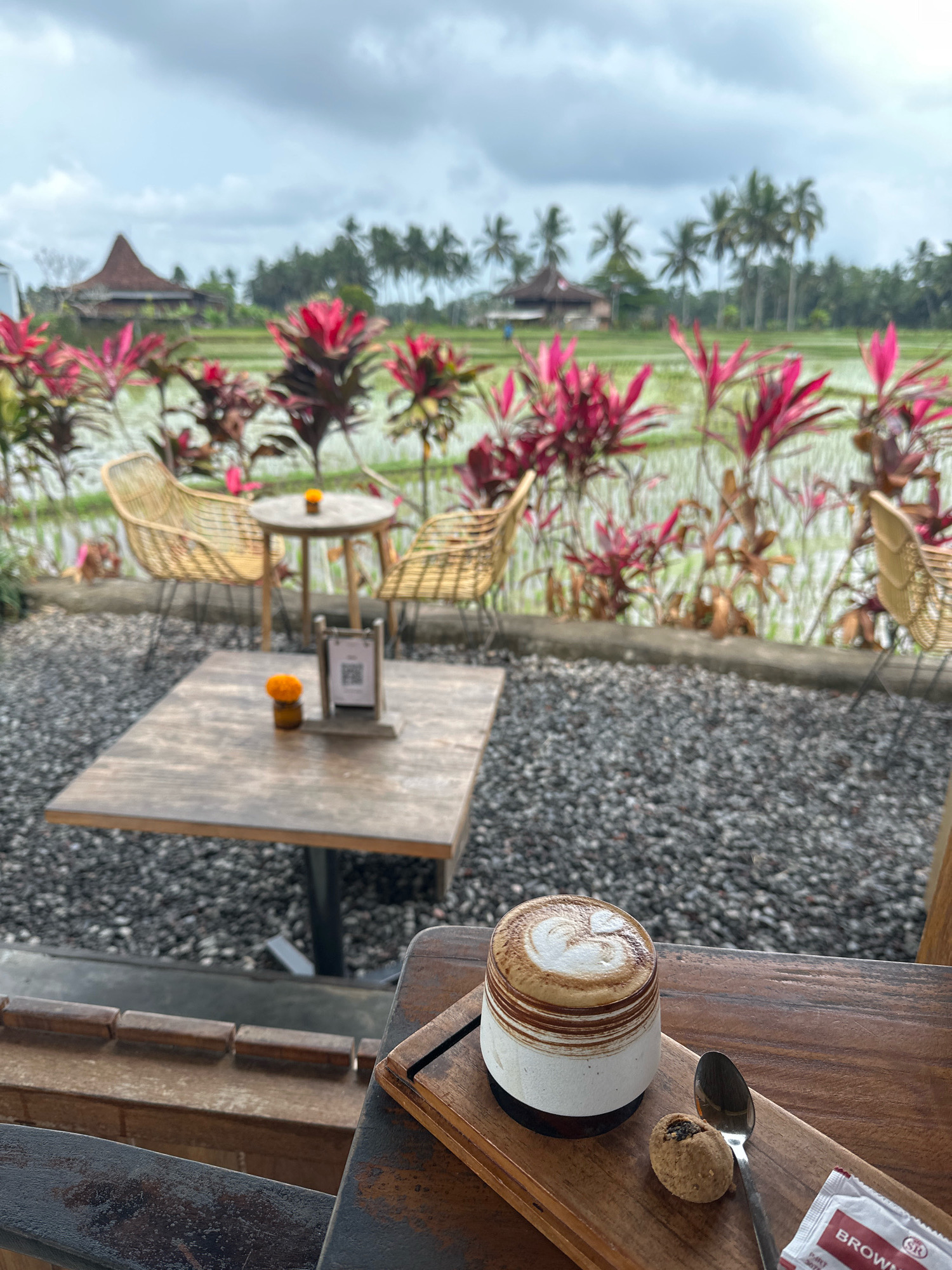 An overhead, medium shot captures a cup of coffee with latte art, a small cookie, a spoon, and a packet of sugar on a wooden tray. The tray sits on a dark wooden table. In the background, a blurred scene shows wicker chairs and tables arranged in front of a lush, green rice paddy field under a cloudy sky. Palm trees and traditional structures are visible in the distance.