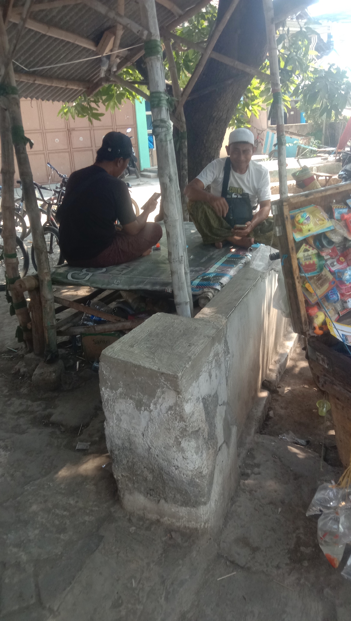 In Ngempit, Indonesia, two men are seated under a makeshift wooden and corrugated metal shelter. The man on the left, wearing a black t-shirt and a dark baseball cap, is looking at his phone while sitting on a raised platform. To his right, another man, dressed in a white t-shirt and a white skullcap, is also holding a phone, in a squatting position on the same platform. He wears a black bag across his chest. The shelter is supported by bamboo poles. In front of the shelter, to the right, is a wooden display case filled with various colorful toys. The ground is dirt and concrete. Bags containing small, orange fish are hanging from a string on the right side of the image.