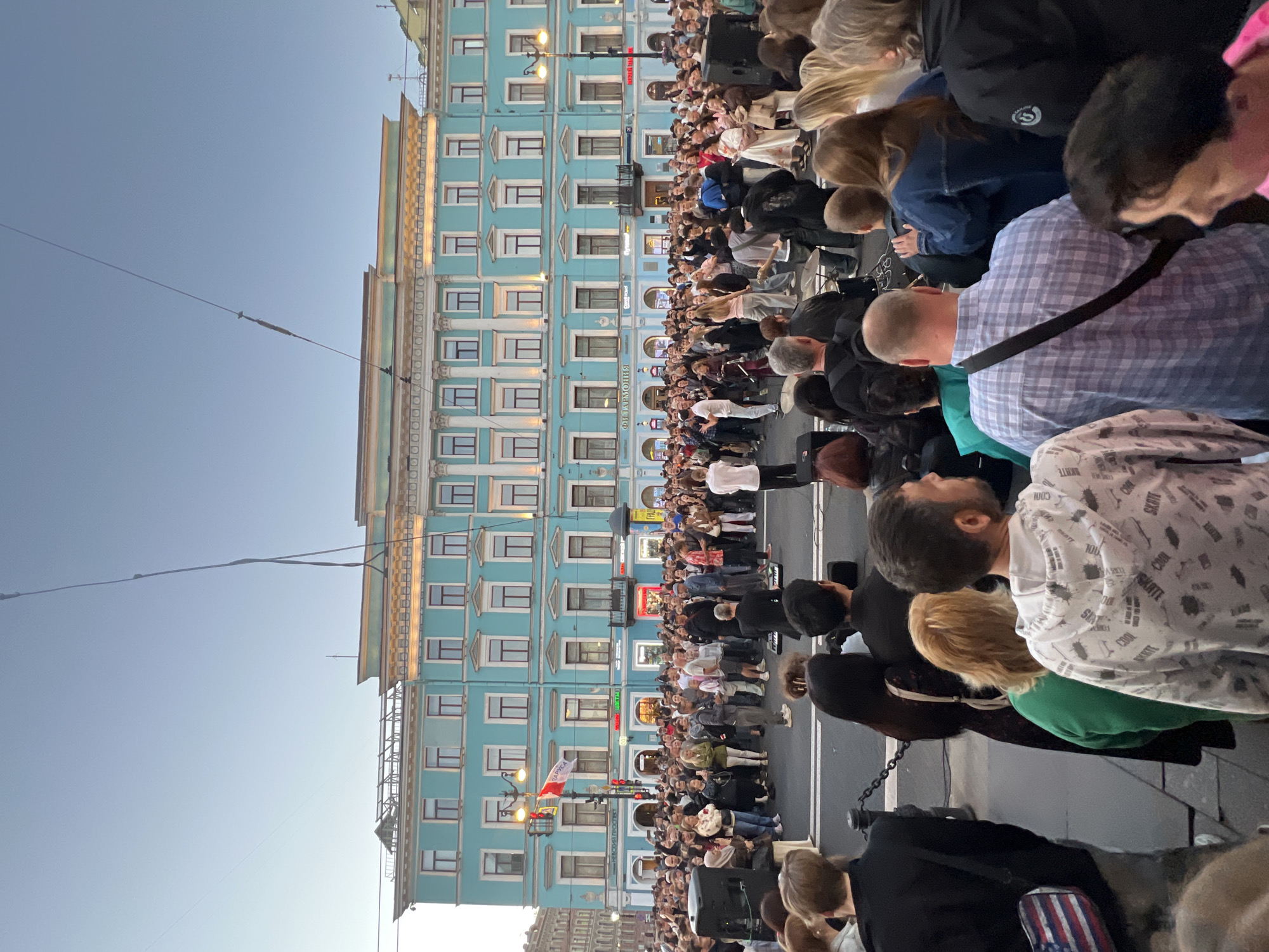 The image captures a street performance in front of a turquoise building in Saint Petersburg, Russia. A crowd of people are gathered to watch a band perform on a temporary stage set up in the street. The building, with its classical architecture, appears to be a public institution, possibly a concert hall or a historical landmark. The street is paved, and there are streetlights and traffic signals visible, indicating an urban setting. The sky is clear and a soft blue, suggesting it is either early morning or late evening. The overall atmosphere is one of a lively public event.