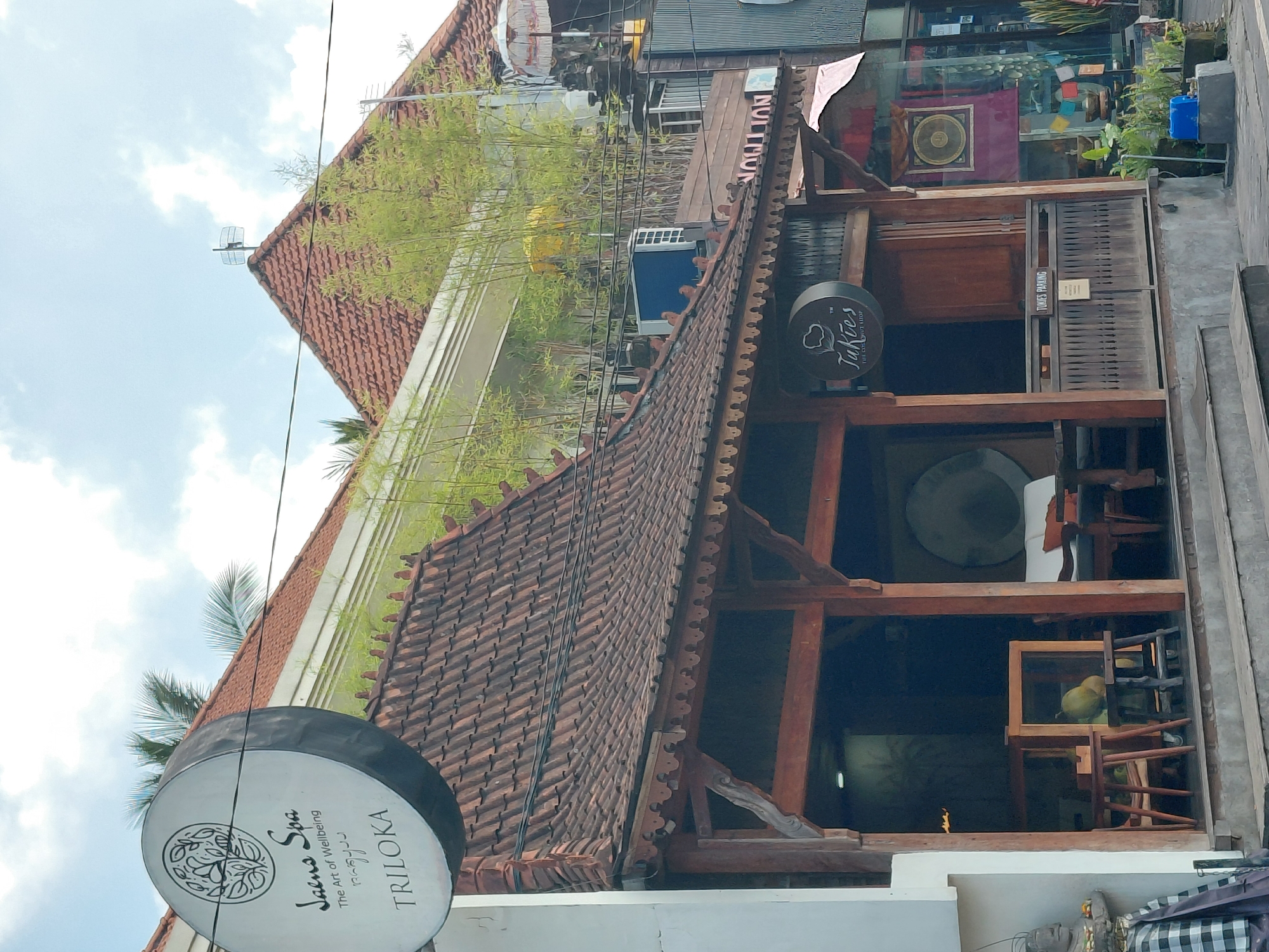 The image depicts a street scene in Ubud District, Indonesia, focusing on the exterior of a building with traditional Balinese architecture. In the foreground, a wooden structure with a tiled roof and decorative eaves shelters an entrance. Inside, furniture and decor are visible, including chairs, tables, and a large circular object, possibly a gong. A sign for "Takies" is prominently displayed. To the left, a circular sign for "Jano Spa - The Art of Wellbeing - Triloka" is attached to a white pillar. The background shows more buildings with red tiled roofs, lush green bamboo plants, and what appears to be an air conditioning unit. A sign with the word "TION" is visible on a building in the distance. The sky is partly cloudy. The scene appears to be a commercial area with shops or businesses.
