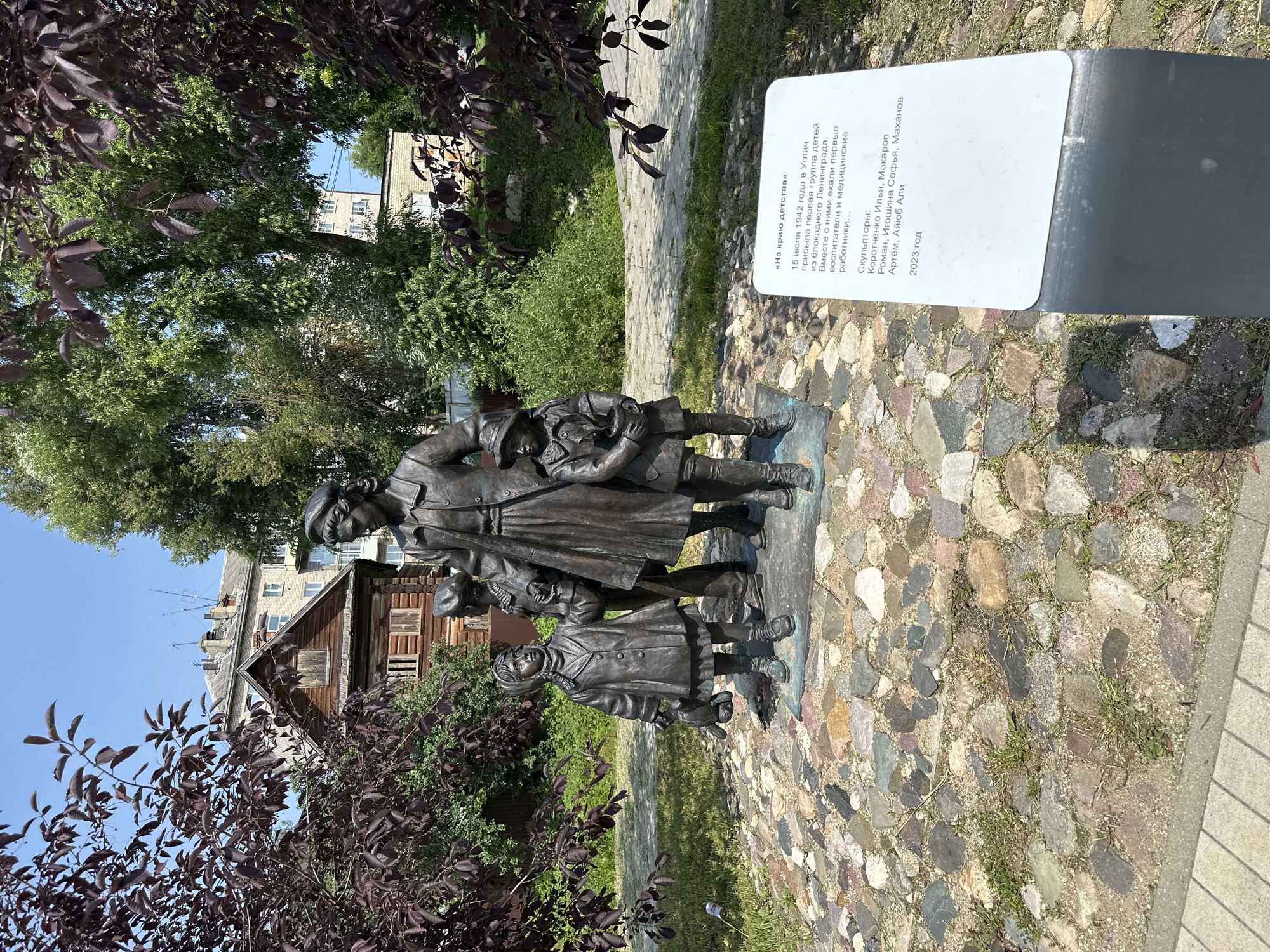 The image shows a bronze sculpture of a woman with three children in an outdoor setting. The sculpture is placed on a base of cobblestones. The woman is wearing a coat and a hat, and she is holding the hands of two children. One child is holding a doll, and the other is holding a plant in a pot. Behind the sculpture, there is a wooden house and a brick building. There are also trees and bushes in the background. A sign with Russian text is visible in the foreground. The sign is likely a plaque or information board related to the sculpture. The scene is lit by sunlight, suggesting it is daytime. The overall environment appears to be a park or a public space. Based on the provided information, the sculpture is located in the city of Terpilovo, Russia.