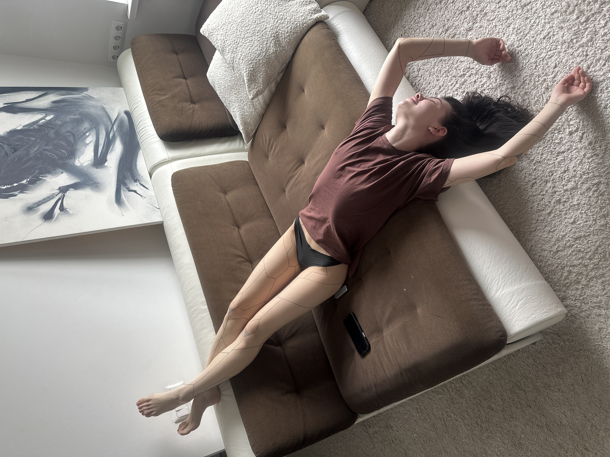 A woman lies on a brown and white sofa, stretching her arms above her head. She wears a brown t-shirt and black underwear. Linear designs are visible on her arms and legs. A black mobile phone rests on the sofa beside her. The sofa has white cushions and a white bolster pillow. An abstract black and white painting is on the wall near the sofa. The floor is covered with a light grey carpet. The location is not specified in the image, nor can be accurately determined.