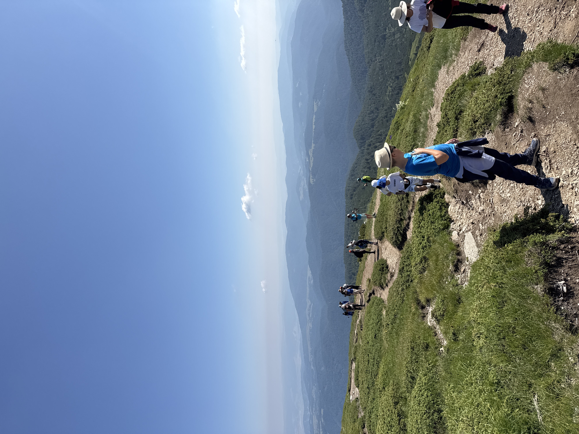 In Panichishte, Bulgaria, a group of hikers is traversing a mountainous landscape under a clear blue sky with sparse clouds. The foreground is dominated by a well-worn dirt path winding through green, low-lying vegetation. Several individuals are visible on the path. Closest to the viewer, a young person wearing a blue t-shirt, dark pants, and a hat is facing away, looking towards the distant mountains. Further up the path, another person in a white t-shirt and hat is seen with their back to the camera. A larger group of hikers is further along the trail, some with backpacks and trekking poles, appearing to be ascending or descending. The background reveals successive layers of rolling, forested mountain ranges fading into a hazy horizon. The overall scene captures a moment of outdoor recreation in a scenic, mountainous environment.