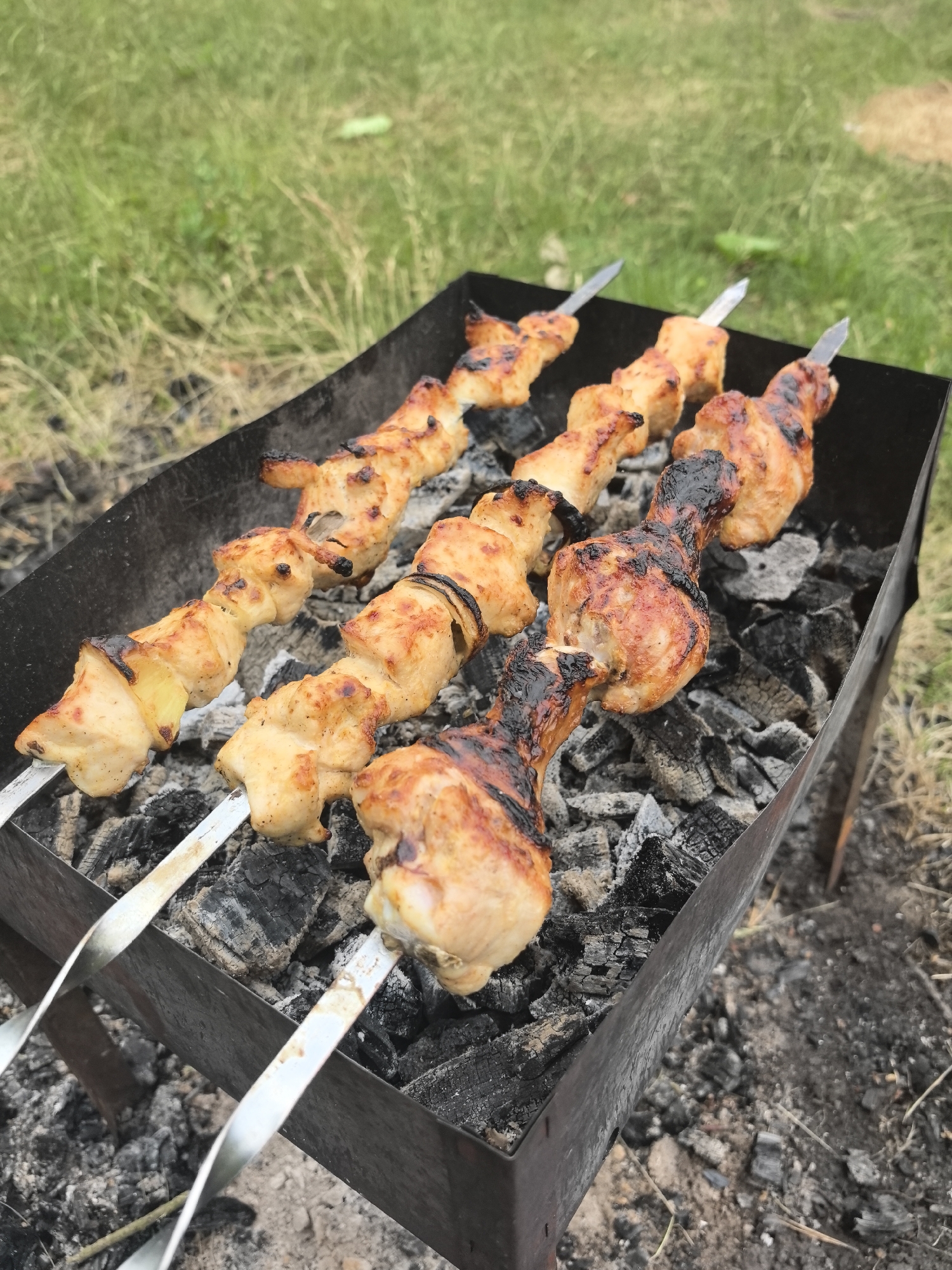 Captured in Парэчча, Belarus, the scene shows a charcoal grill with three skewers laid across it. The skewers hold varying types of poultry, including cut pieces of chicken and chicken drumsticks. The grill is set on the ground near grassy vegetation. The skewers are made of metal. The charcoal in the grill is ashen and burnt.