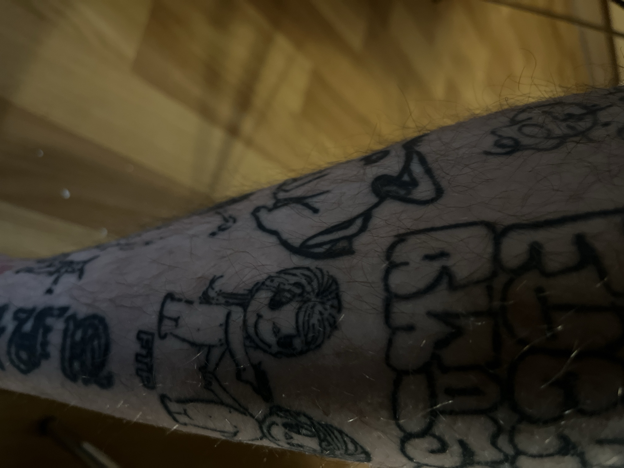 The image is a close-up, full-body shot of a person's leg adorned with multiple black ink tattoos. The tattoos are varied in style, including graffiti-style lettering, cartoonish figures, and possibly abstract designs.
In the upper portion of the visible leg, there is a tattoo that appears to be graffiti lettering, rendered in a bubble font. Below this, a tattoo depicts a cartoon dog. Further down, a tattoo shows two stylized figures. The figure on the left has a unique hairstyle and is holding what appears to be a gun, pointing it towards the figure on the right. The figure on the right is also a stylized character. Below these figures, the letters "FTP" are tattooed. At the bottom of the frame, more tattoos are visible, including what looks like a circular design and another abstract or figurative tattoo. The leg itself is hairy, and the background appears to be a wooden floor with a warm, yellow-brown hue, suggesting an indoor setting. The lighting in the image is somewhat dim, casting shadows on the floor. There are no identifiable landmarks or context to determine a specific city or location.