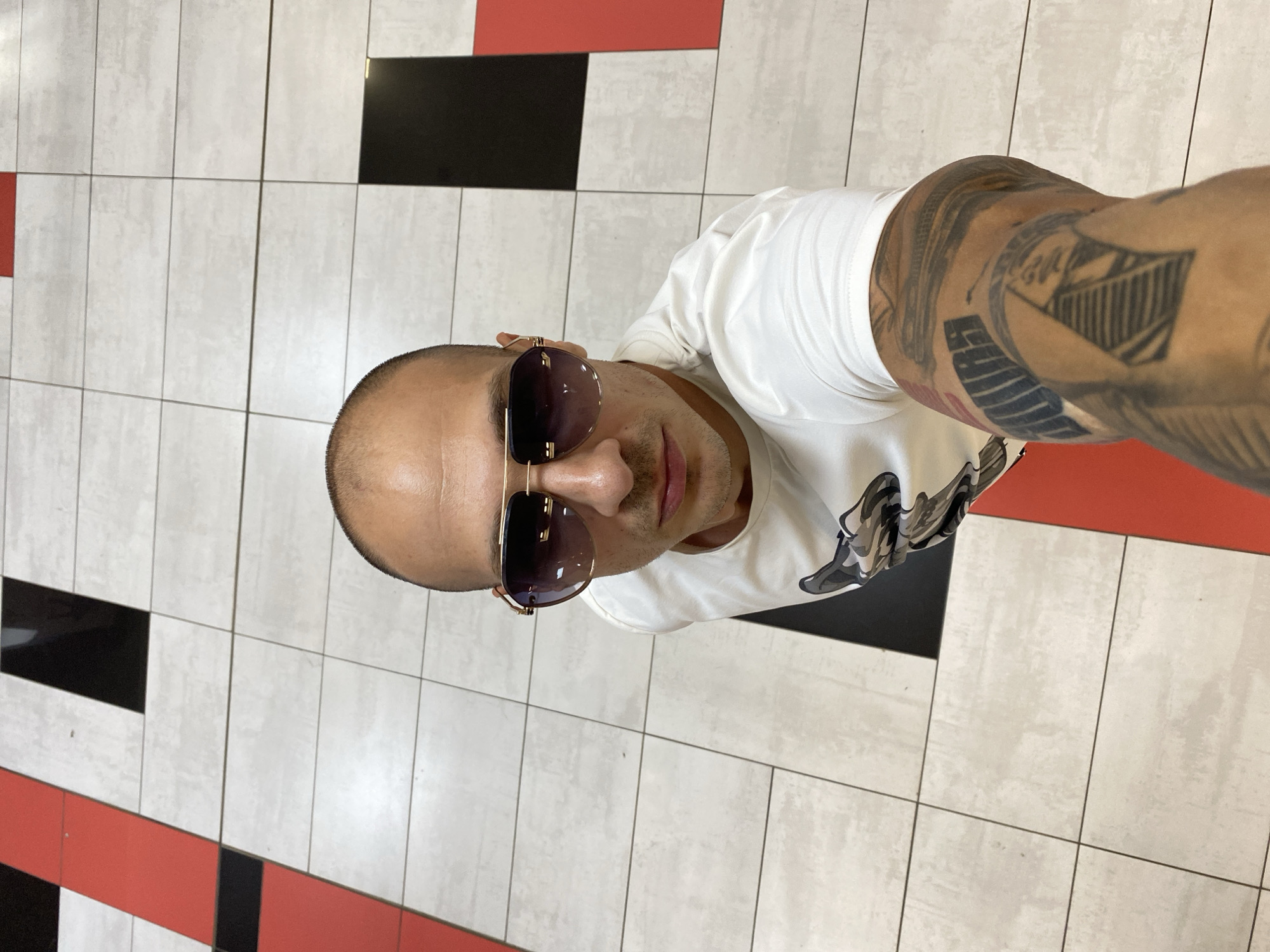 A man wearing sunglasses and a white t-shirt with a camouflage print is taking a selfie in what appears to be a public indoor space with a patterned tiled floor. The floor is made of large, square tiles in shades of light gray, with some sections featuring alternating black and red rectangular accents. The man's arm, visible in the foreground, is covered in tattoos, including what appears to be a pyramid design and text. The lighting suggests an overhead light source. The location is identified as the City of Pleven, Bulgaria.