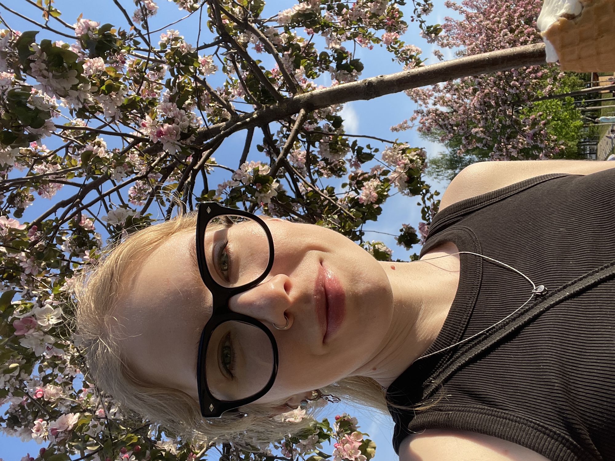 The image is a selfie of a person with fair skin and blonde hair, wearing black-framed glasses and a black tank top. They have a nose ring and are wearing a delicate silver necklace. The background features flowering trees with pink and white blossoms against a blue sky with some clouds. Part of an ice cream cone is visible in the top right corner. There are no identifying features to determine the exact location in Izhevsk, Russia.