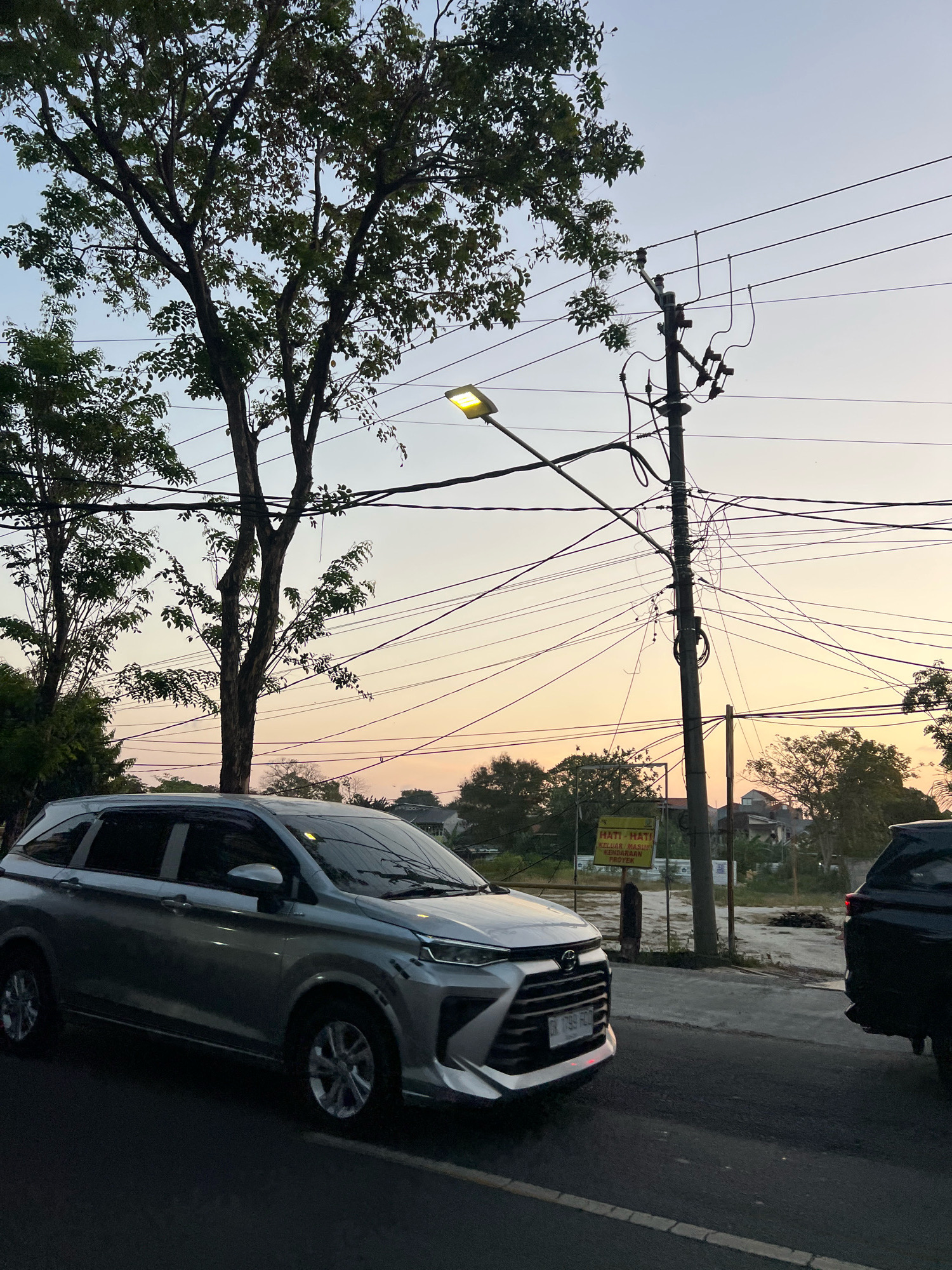 The image captures a street scene in Kuta, Indonesia, during the late afternoon or early evening. The sky is a soft gradient of pale blue and warm orange, indicating a sunset. A silver minivan is parked on the left side of the road, facing right. To the right of the minivan, a black SUV is partially visible, also on the road.
A prominent utility pole stands on the right side of the frame, heavily laden with a complex network of electrical wires. A street lamp is attached to the pole on the left, emitting a yellow light. Trees with full foliage line the left side of the street, their branches reaching towards the sky. In the background, there are buildings and more trees, with a yellow sign visible on a post that reads "HATI HATI KELUAR MASUK KENDARAAN PROYEK". This translates to "BE CAREFUL ENTRY AND EXIT VEHICLES PROJECT". The road surface is asphalt, with a white line marking the edge of the lane. The overall impression is of a typical urban street in a tropical climate at dusk.