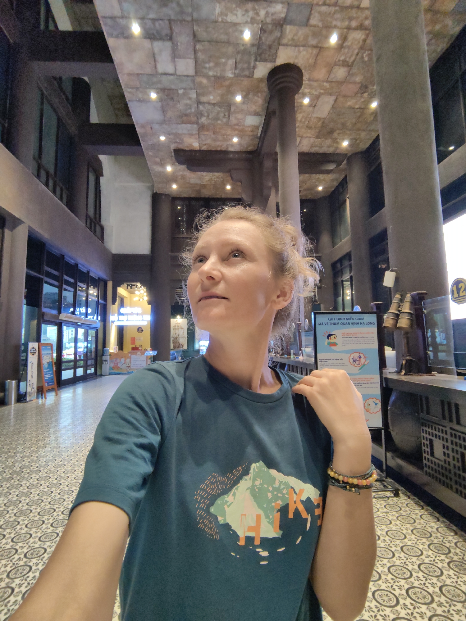 A fair-skinned woman with blonde, curly hair is standing indoors, looking up and to the left. She is wearing a teal t-shirt with a mountain graphic and the word "HIKE" printed on it. She is also wearing multiple beaded bracelets on her right wrist. The background reveals a spacious interior, with patterned tile flooring and tall, dark pillars supporting a decorated ceiling with recessed lighting. Glass doors are visible in the distance, leading to an exterior area. A sign with Vietnamese writing and illustrations is on the right side of the frame. The photo was taken in Bai Chay Ward, Vietnam.
