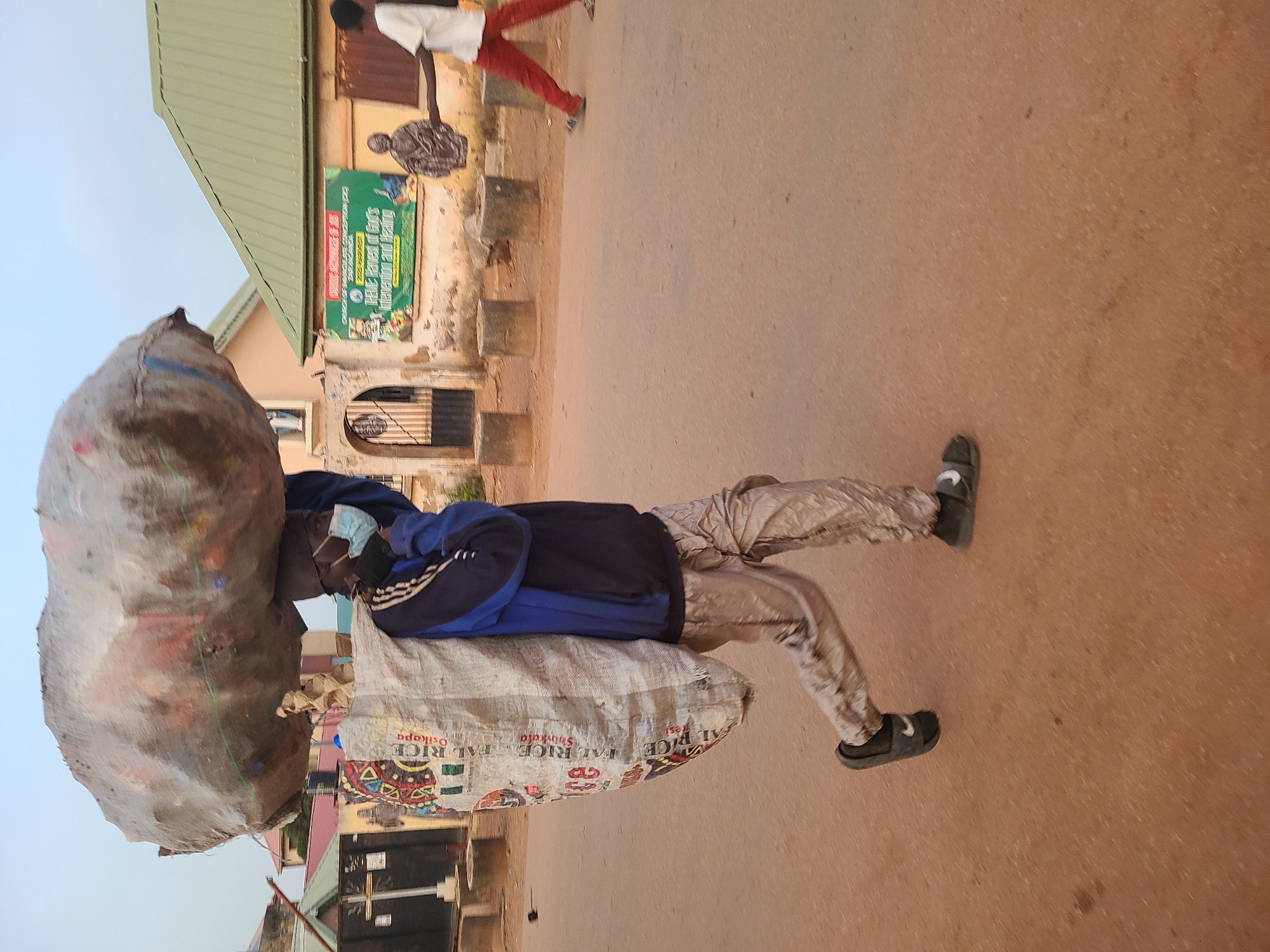 An African man in a blue and black jacket, and light-colored camouflage pants, and black slide sandals walks down a paved street carrying two large bags on his head and shoulder. He wears a medical face mask.
The bags obscure a building with a green roof, a green and white sign that reads "CHURCH OF IMMACULATE CONCEPTION (CGC) 2025 Harvest THEVE Harvest of God's Intervention and Healing", and a fenced entryway.
Another person in a white shirt and red pants is further down the street, walking in the opposite direction.
The words "AL RICE" and "Osikapa" are visible on one of the bags.
The image location is Jos, Nigeria.