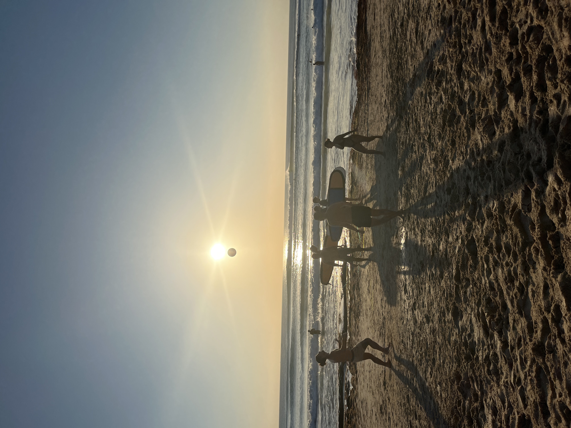 A group of silhouetted individuals are playing a game of volleyball on the sandy beach at Kuta Selatan, Indonesia, during sunset. The sun is low in the sky, casting a warm glow and creating long shadows on the sand. The ocean stretches out behind them, with gentle waves rolling onto the shore and a few surfers visible in the distance. Two surfboards are placed on the sand near the group. The sky is a clear, gradient blue, transitioning to a golden hue near the horizon. The foreground of the image is dominated by the textured sand of the beach.