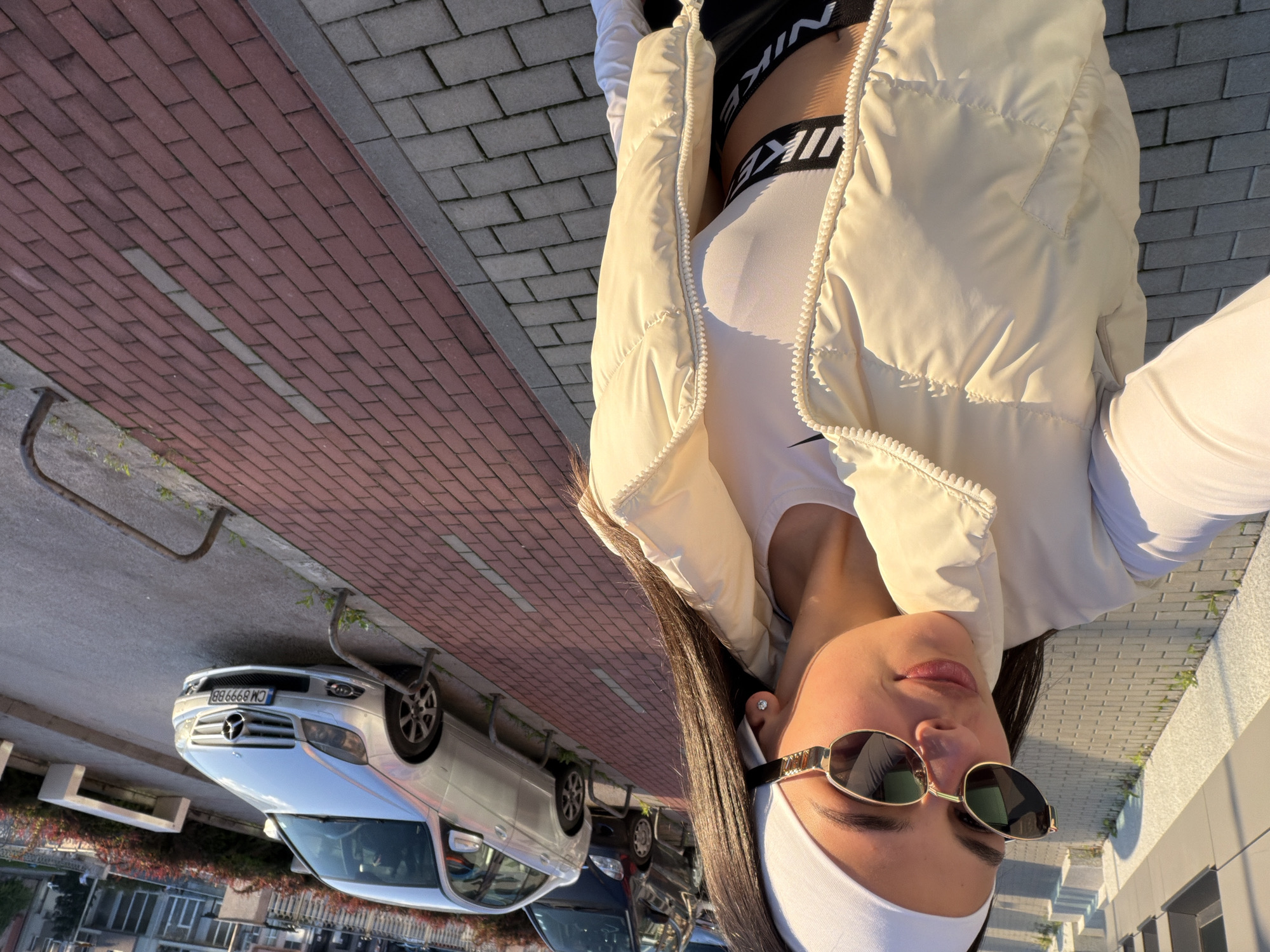 A street-level photograph shows a young woman, wearing sunglasses, a white sports headband, a white sleeveless shirt, a black sports bra with "NIKE" prominently displayed, and a white puffer jacket, standing on a brick sidewalk. She is facing the camera. A silver Mercedes-Benz car is parked along the street. Several buildings are visible in the background. The image was taken in Gabrovo, Bulgaria based on the license plate on the vehicle.