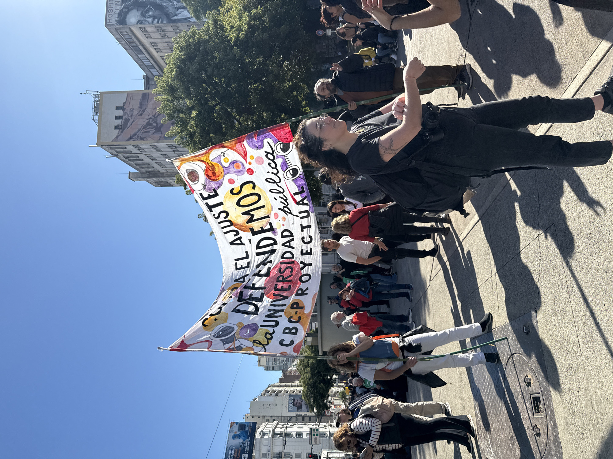 The image shows a protest march in what appears to be Buenos Aires, Argentina. A large banner reads, "Defendamos El Ajuste a la Universidad Publica, CBC-Proyectual." A woman in black carries one end of the banner. The crowd is walking on a sidewalk in an urban environment. Buildings, including one with a mural, line the street. The sky is clear and blue. The perspective is tilted and a sewer cap is visible in the foreground.