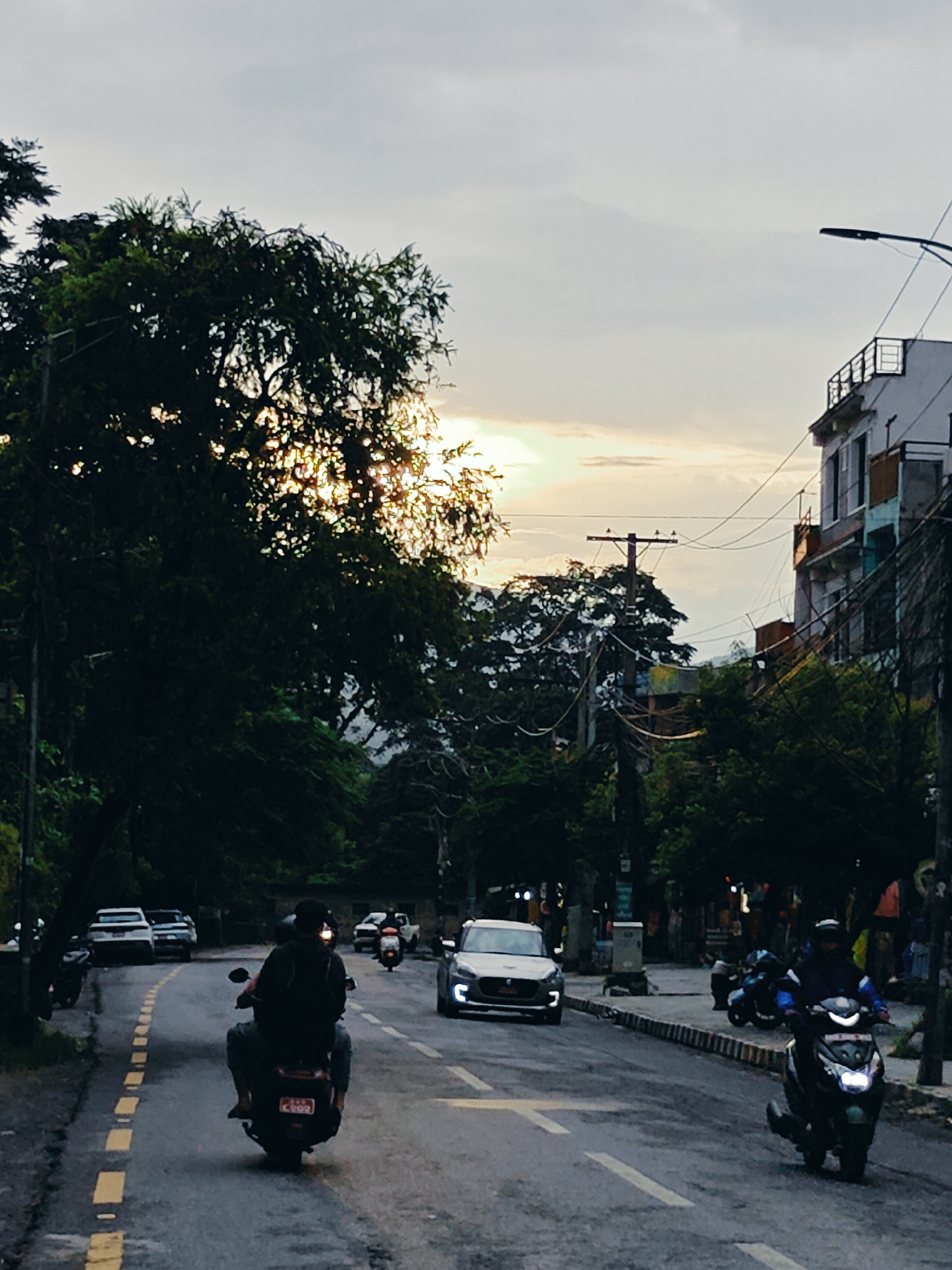 The image depicts a street scene in Pokhara, Nepal, during twilight. A road with yellow and white dashed lines runs through the center, with vehicles and pedestrians present. On the left side of the road, a person on a scooter is seen from behind, heading away from the viewer. Further down the road, several cars and motorcycles are visible. On the right side of the road, a scooter with its headlights on is moving towards the viewer. Buildings line the right side of the street, with trees and foliage forming a canopy on the left. The sky is overcast with hints of sunlight breaking through the clouds. The overall atmosphere is that of a busy urban environment at the end of the day.