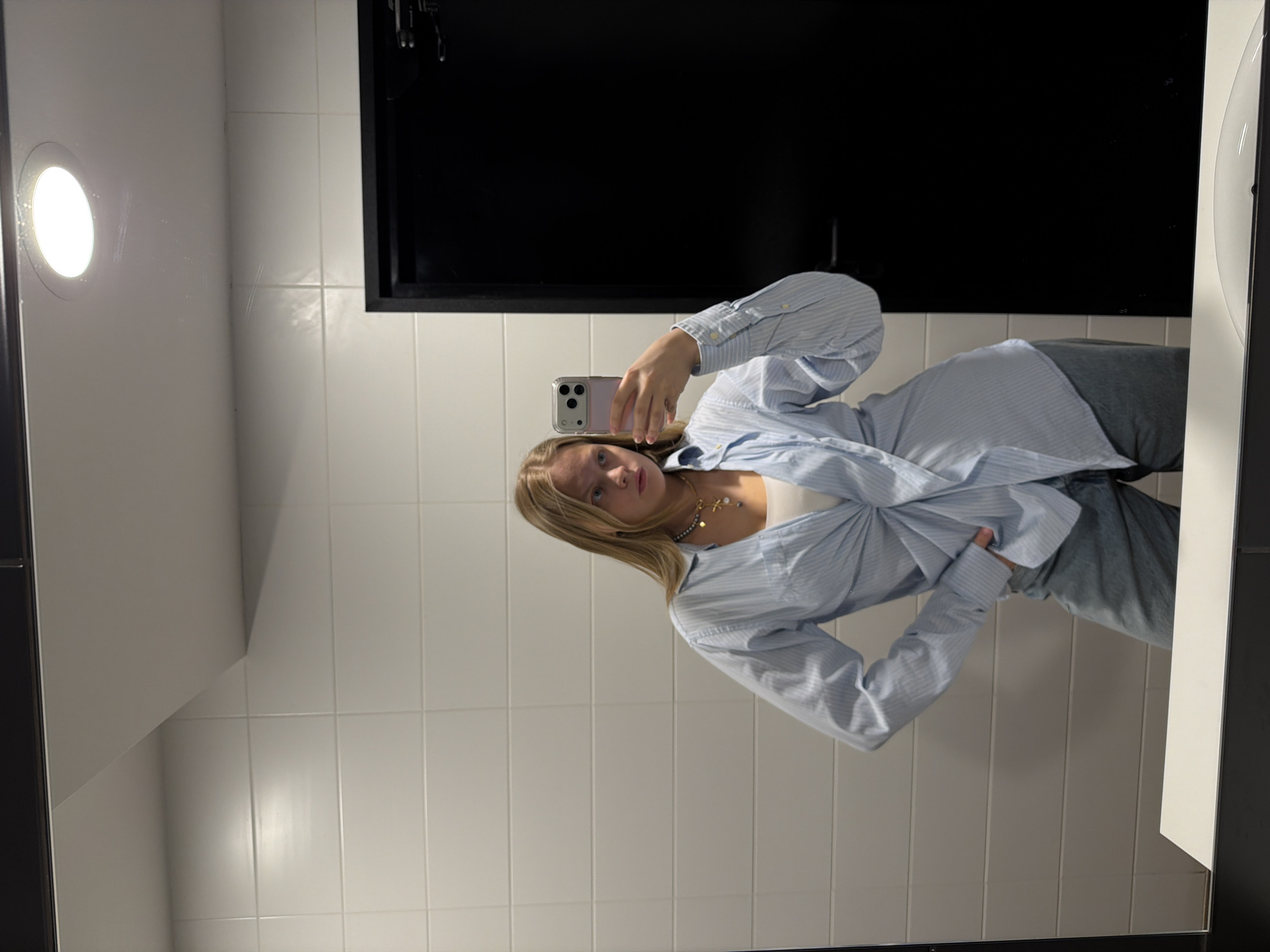 A young woman is taking a selfie in a bathroom, possibly in a public setting. The bathroom has white tiled walls, a black cabinet above the sink, and a round ceiling light. The woman is wearing a light blue striped shirt over a white top and gray pants. She is holding a smartphone in her left hand, using it to capture her reflection in the mirror. Her posture suggests she is posing for the photo. The location cannot be confirmed to be in Izhevsk, Russia.
