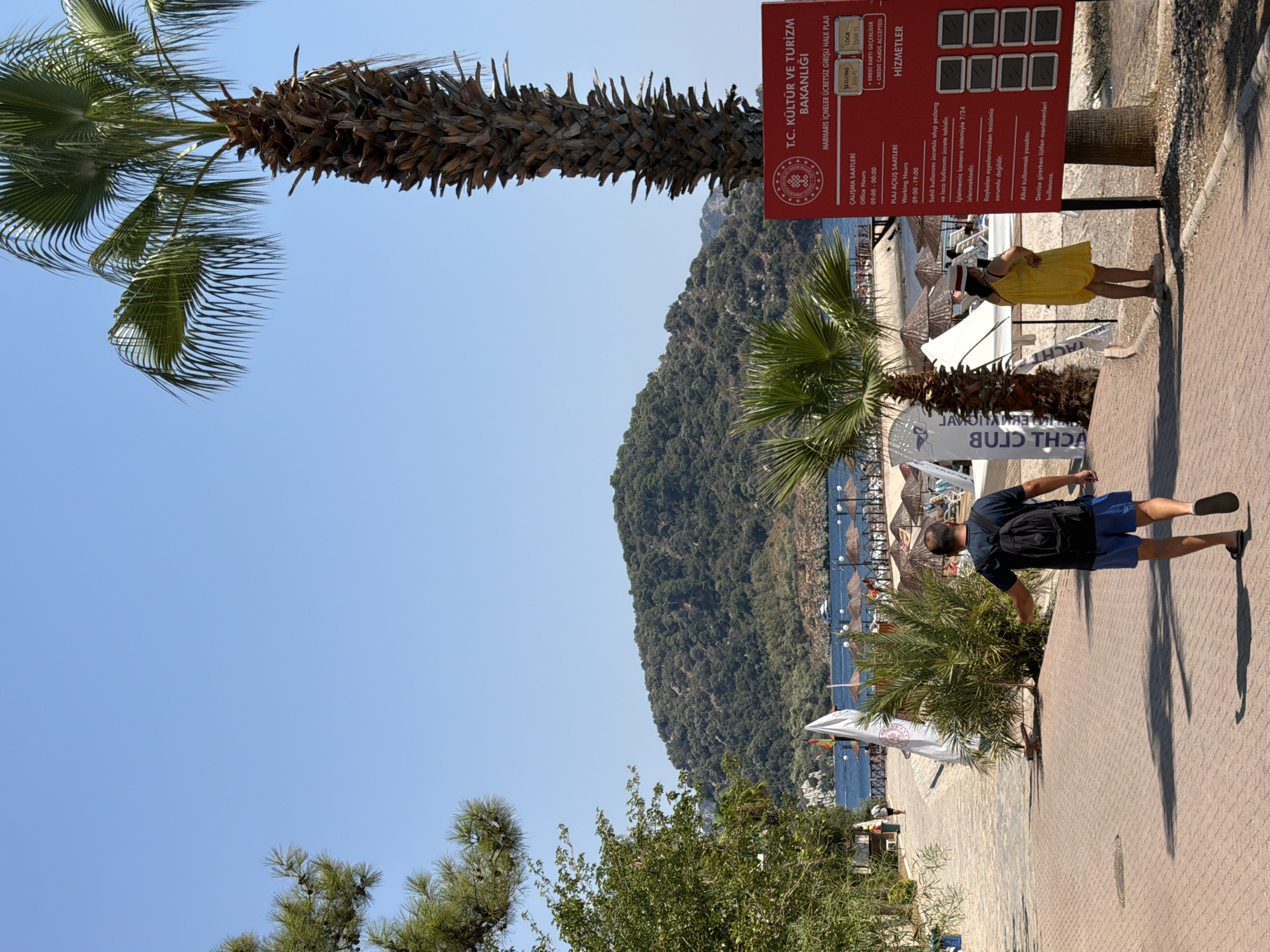 This is an eye-level, medium shot of a public beach area in Marmaris, Turkey, on a clear, sunny day. A man wearing a dark blue t-shirt, blue shorts, and black sandals is walking away from the camera on a paved walkway. He has a black backpack on. To his right, a woman in a yellow dress and a white hat stands with her hands on her hips, facing away from the camera and to her left.
In the foreground, a large palm tree with a rough, textured trunk is on the right side of the image. To the left of the walkway, there are more palm trees and green foliage. A large red sign with white text is posted near the palm tree, with the heading "T.C. KÜLTÜR VE TURİZM BAKANLIĞI" which translates to "Republic of Turkey Ministry of Culture and Tourism". Below this, it states "MARMARİS İÇMELER ÜCRETSİZ GİRİŞ HALK PLAJI" which means "Marmaris İçmeler Free Entrance Public Beach". The sign also details office hours, working hours, and services.
In the background, a sandy beach is visible with rows of straw umbrellas and lounge chairs. People are scattered on the beach and near the water. The sea is a deep blue, and beyond the beach, a lush green, tree-covered hill dominates the landscape under a clear blue sky. A white boat is anchored in the water near the hill. The walkway is made of red brick pavers with shadows cast by the trees and people.