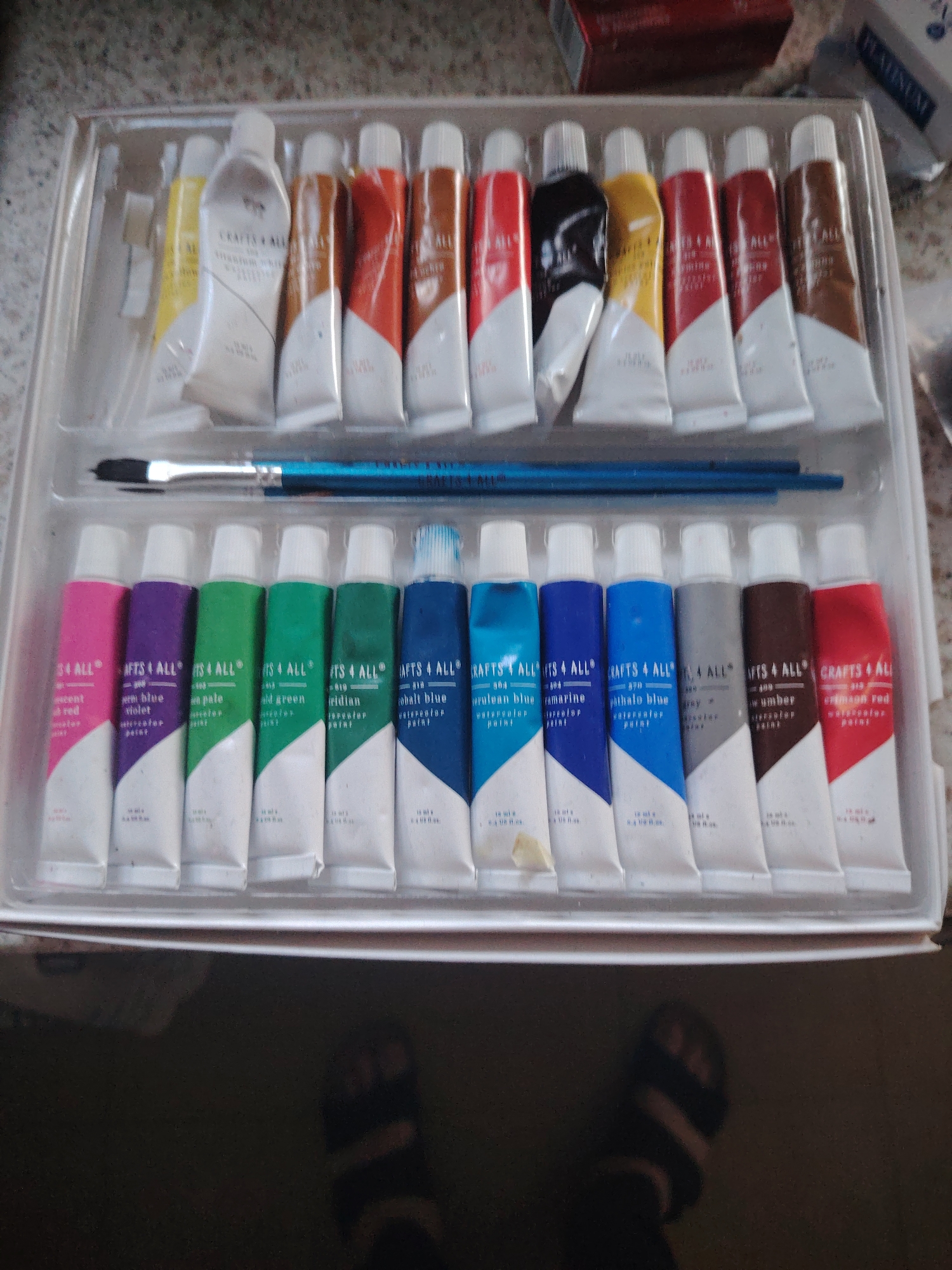 A person's feet wearing black and white striped socks and black sandals are visible at the bottom of the image. Above the feet is a clear plastic tray containing tubes of watercolor paint and a paintbrush. The tray holds two rows of paint tubes. The top row contains tubes of white, yellow, brown, orange, and red paint. The bottom row contains tubes of pink, purple, green, blue, grey, brown, and red paint. The tubes are labeled with "CRAFTS 4 ALL" and the color name. The background is blurred and appears to be a countertop. There is no indication of the city of Huntingdonshire, United Kingdom in the image.