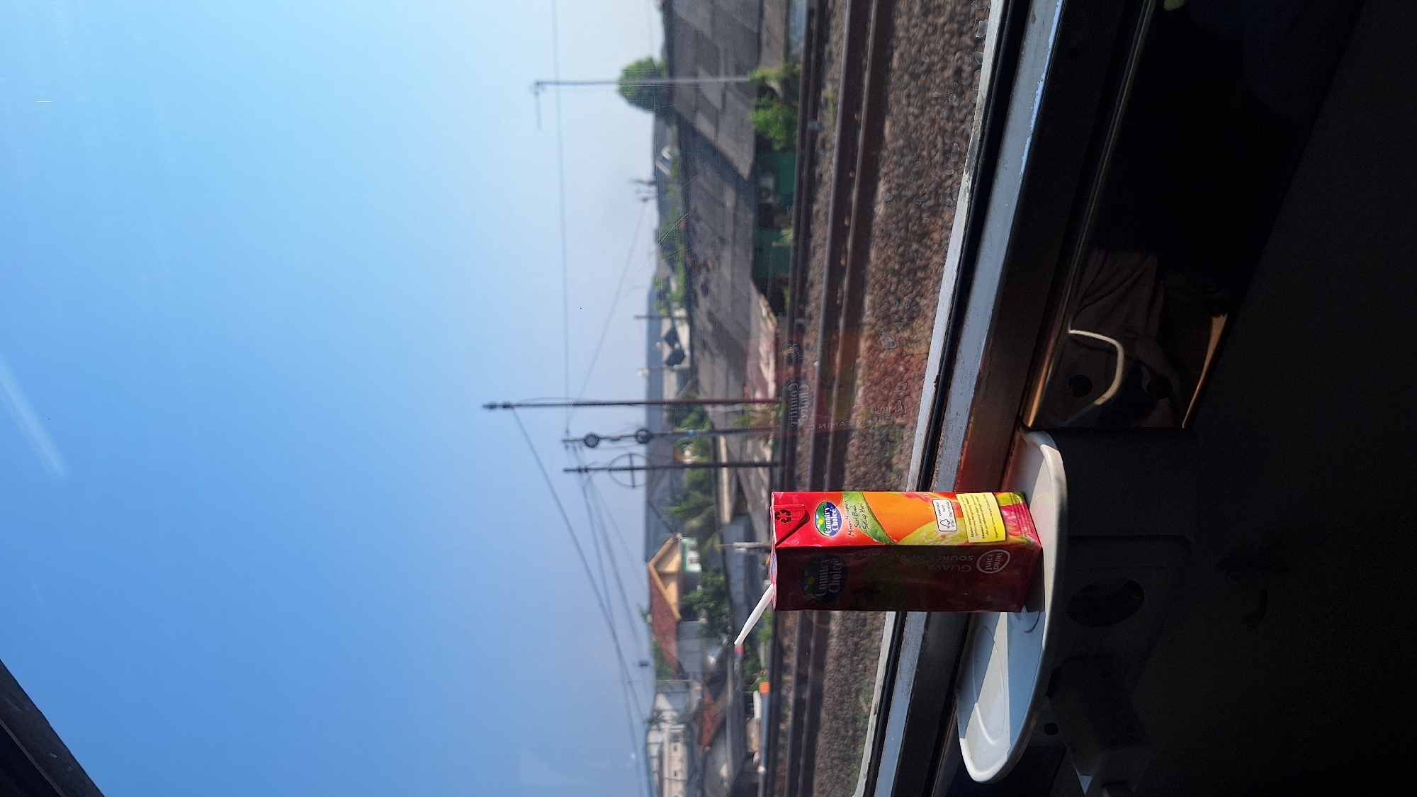 A juice box and a straw sit on a tray table in front of a train window, looking out at a sunny landscape of houses and train tracks in Waringinjaya, Indonesia. The sky is clear and blue. The juice box is red and features the "Country Choice" logo. The houses in the distance have tiled roofs and are surrounded by trees. Power lines and utility poles are visible in the background. The train tracks are in the foreground, with gravel and ballast surrounding them. The interior of the train car is dark, with the window providing the primary light source.