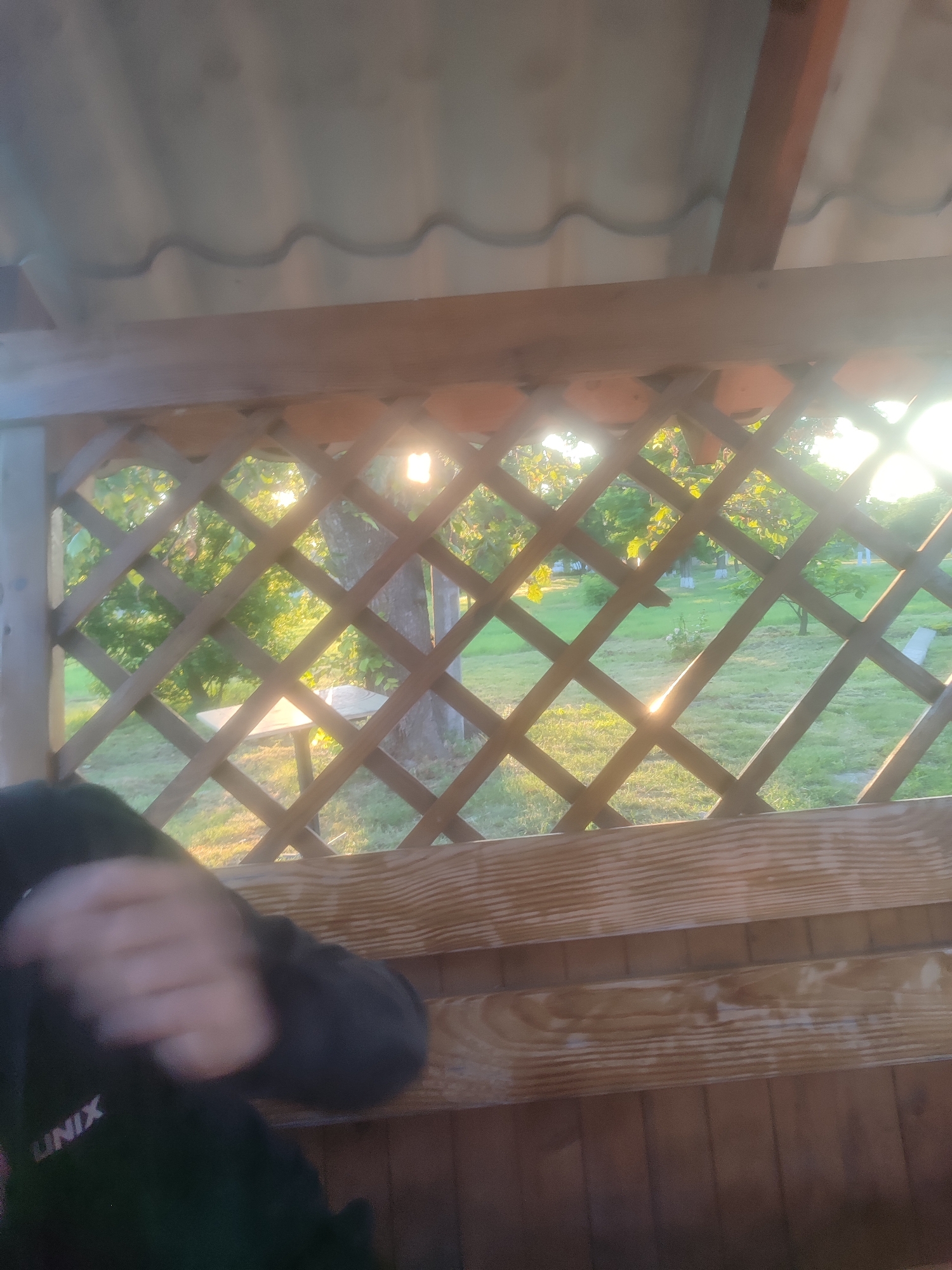 The image, taken from inside a wooden gazebo, looks out onto a grassy yard bathed in the warm light of the setting sun. A lattice pattern of wooden beams divides the view, creating a diamond-shaped frame for the outdoor scene. In the foreground, a dark-colored hooded garment is visible, with the text "UNIX" subtly imprinted on the sleeve. The sun's rays cast bright flares and highlights through the wooden lattice and onto the surrounding trees and grass. The overall impression is one of a peaceful, possibly late afternoon or early evening setting in an outdoor recreational area.