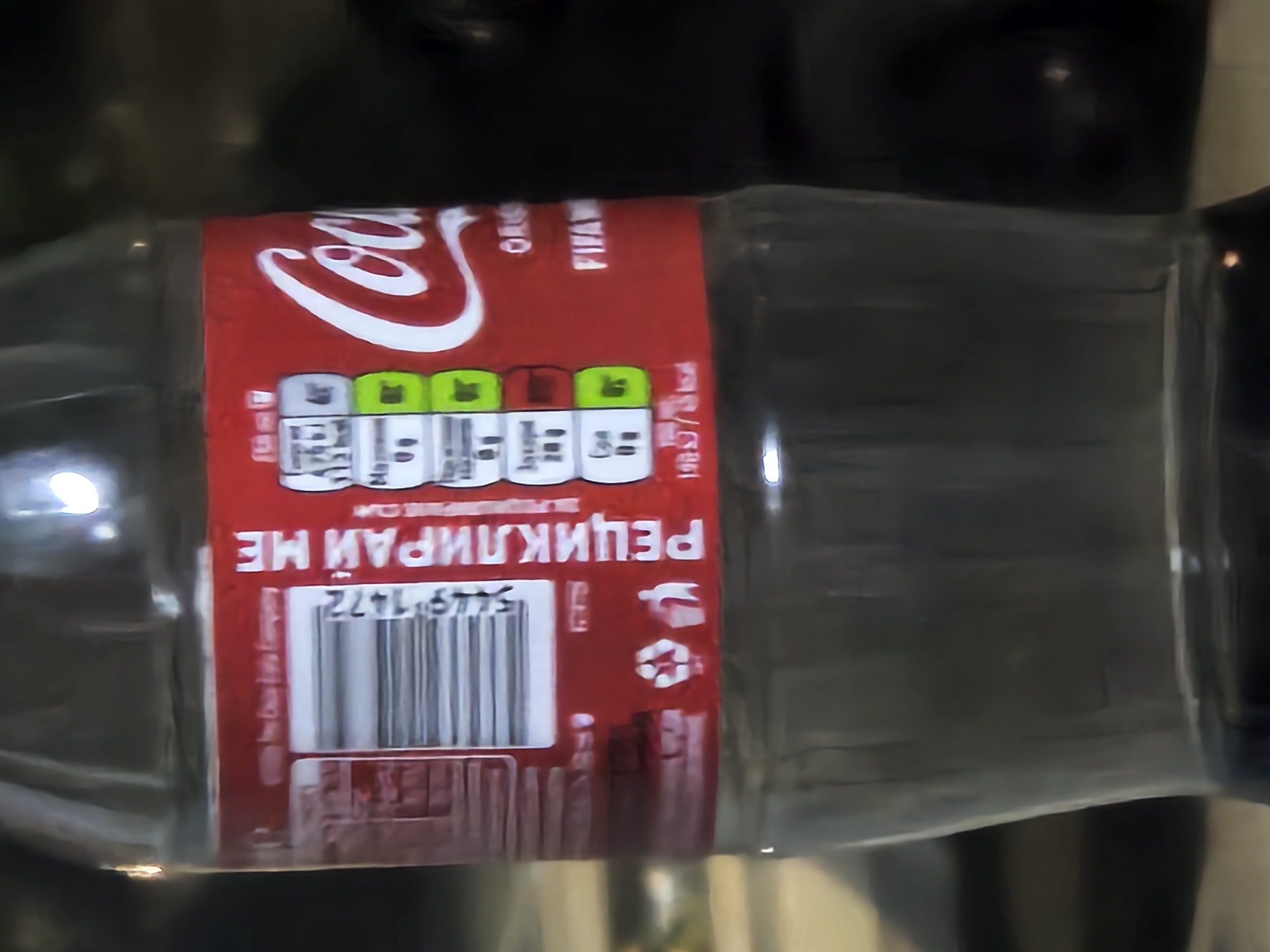 A close-up, low-angle, medium shot captures a portion of a clear plastic Coca-Cola bottle. The label is predominantly red with white text and graphics. On the left side of the label is a barcode and nutritional information presented in a series of colored boxes (white, green, and orange). To the right of this, in large white lettering, is the Coca-Cola logo. Below the logo, the text "ORIGINAL TASTE" and "FIFAR" are visible. In the lower section of the label, there are recycling symbols. The bottle is partially filled with a dark liquid. The background is blurred, appearing to be a dark, possibly indoor environment with some subtle lighting. The specific location in the city of Biaga, Bulgaria cannot be determined from the visual information provided.
