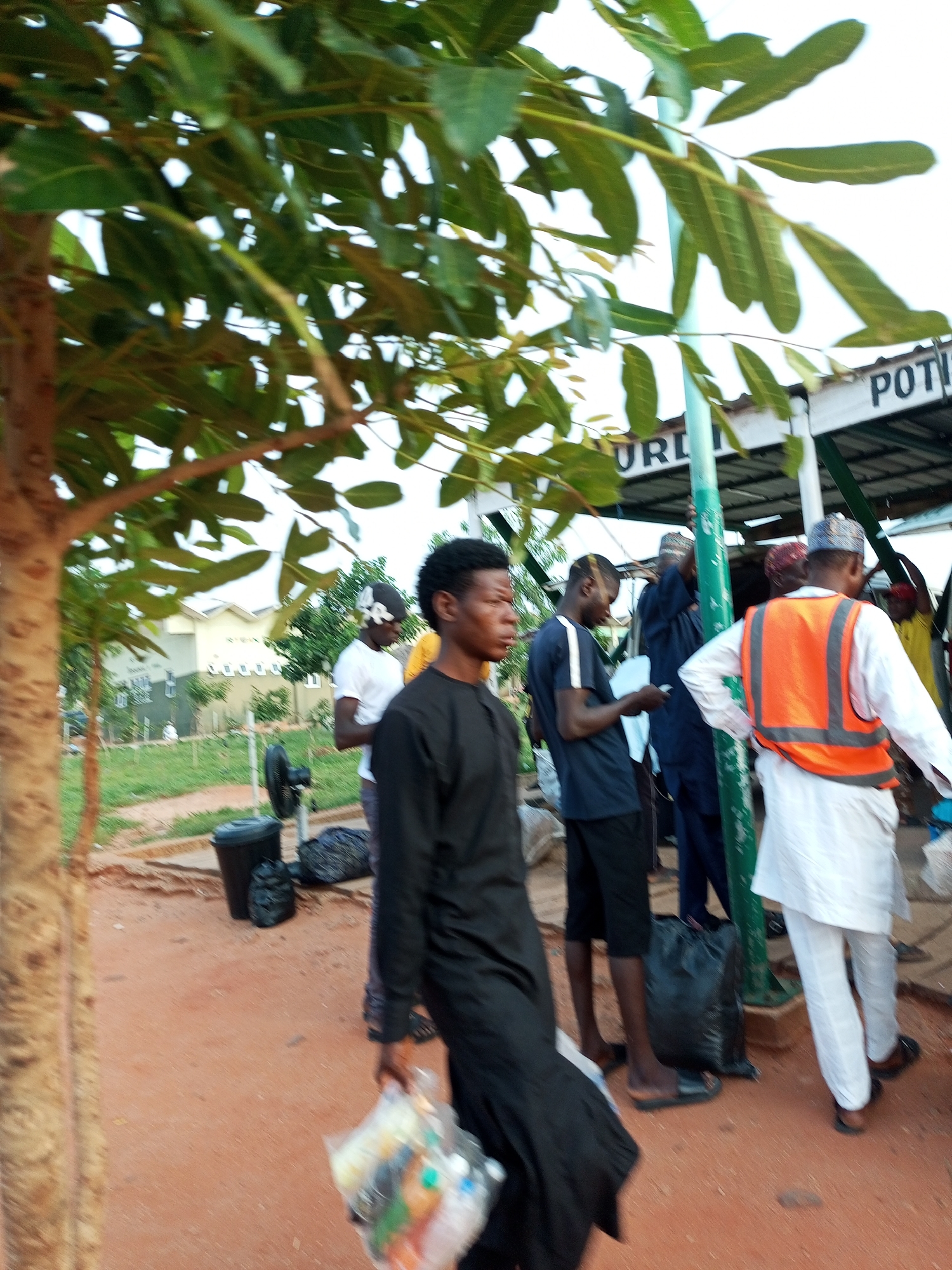 The image depicts an outdoor scene with several people. In the foreground, a young man wearing a black outfit walks from left to right, carrying a clear plastic bag filled with items. To his right, another young man in a dark blue t-shirt and black shorts is standing, holding a piece of paper. Behind him, a man in a dark blue robe is also visible. Further to the right, a person wearing a white outfit and an orange safety vest stands facing away from the camera. Another individual in a white shirt and a patterned head covering is seen in the mid-ground.
The environment appears to be a dusty, reddish-brown outdoor area, possibly a street or marketplace, with some greenery and trees. In the background, a building with a white facade and windows can be seen. There are also signs visible, with partial words like "Poti" and "UPL". A black trash bin and a portable fan are positioned on the ground near the people. The overall lighting suggests it is daytime, possibly overcast.