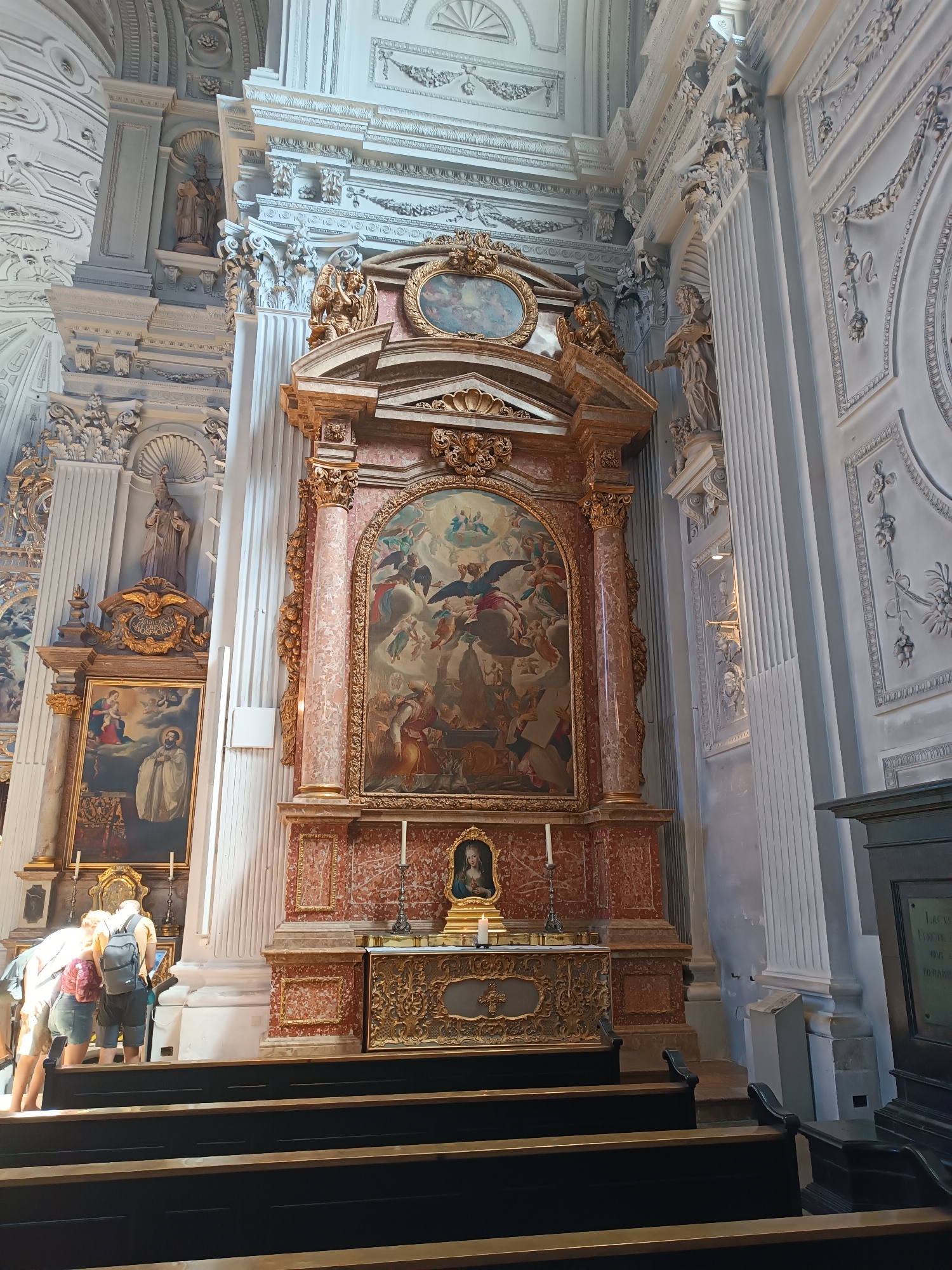 The image depicts the interior of a grand church, specifically the Frauenkirche in Munich, Germany. In the foreground, several dark wooden pews are visible, with the backs of three individuals in casual attire facing away from the camera, looking towards an altar area. The individuals are a woman with blonde hair wearing a pink and white patterned top and denim shorts, another woman with blonde hair in a backpack wearing a white top and grey shorts, and a person wearing a dark blue shirt and grey shorts.
The altar area is ornate, featuring a large, framed painting depicting a religious scene with numerous figures and angelic beings. This central artwork is flanked by grand, marble-patterned columns and ornate gold-colored architectural elements. Above the main painting is a smaller oval frame containing another religious artwork. To the left of the main altar, another framed painting of a religious figure is visible. The walls and ceiling of the church are elaborately decorated with intricate stucco work and moldings, indicative of Baroque architecture. The lighting appears to be natural, with some artificial light sources present, creating a mix of bright and shadowed areas.